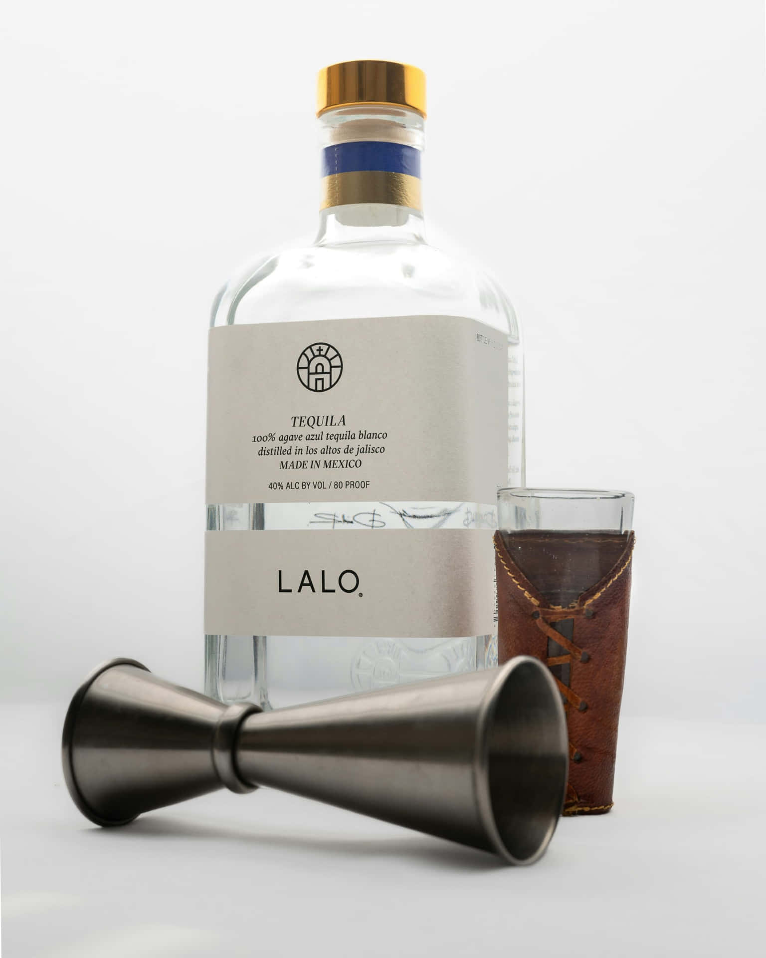  壁紙 — 一個透明嘅Lalo龍舌蘭酒樽，金色樽蓋同簡約標籤，旁邊放咗一個皮革包裹嘅杯同金屬量酒器喺白色表面上。