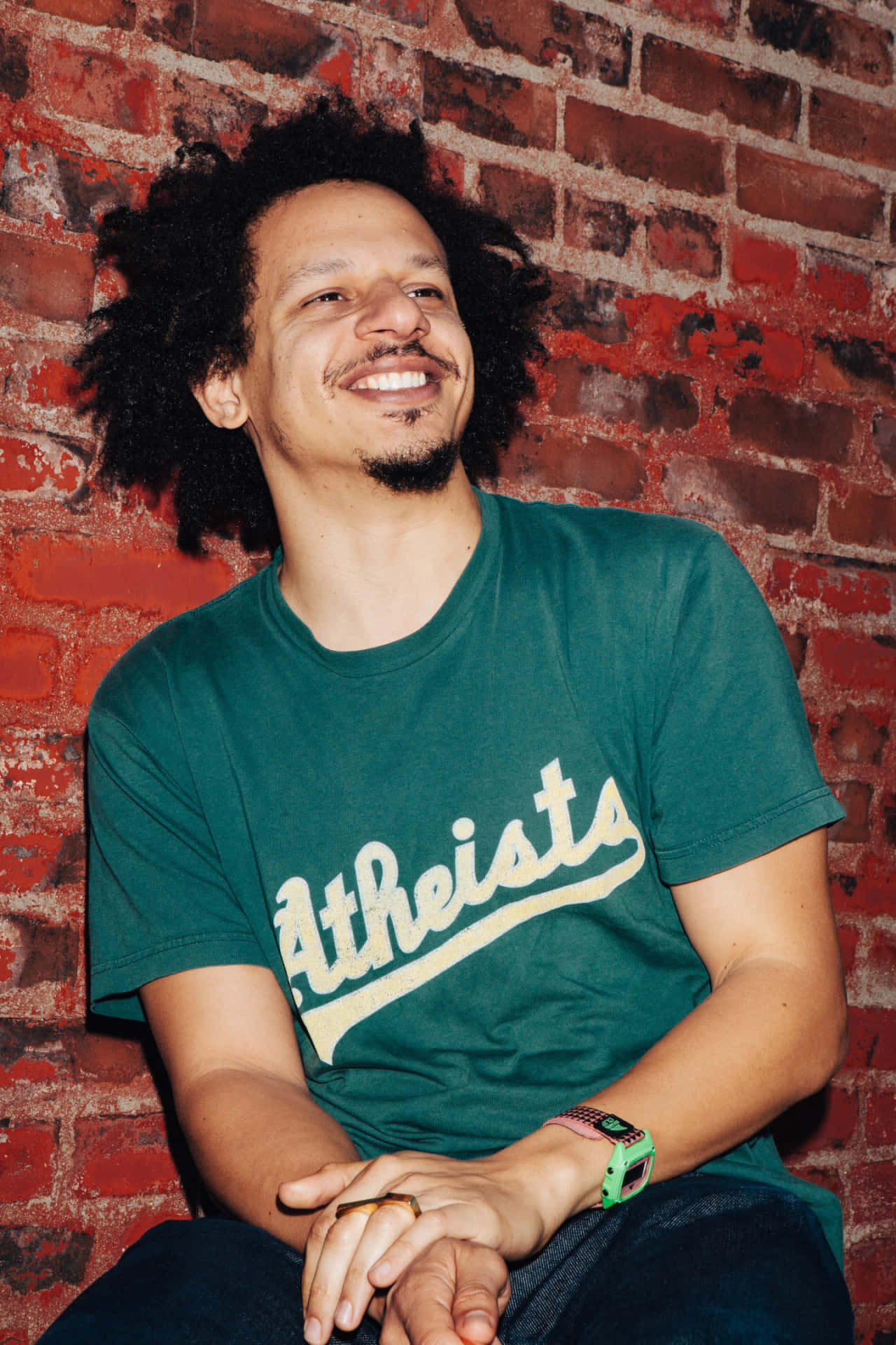  壁紙 — 一個微笑的捲髮男人靠著紅磚牆坐著，穿著綠色'Atheists'T恤。