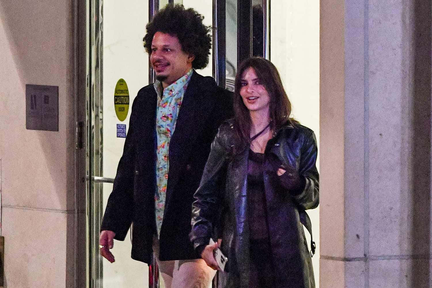  壁紙 — 一個微笑的捲髮男子和一個穿皮褸的女子在夜晚的人造燈光下離開建築物。