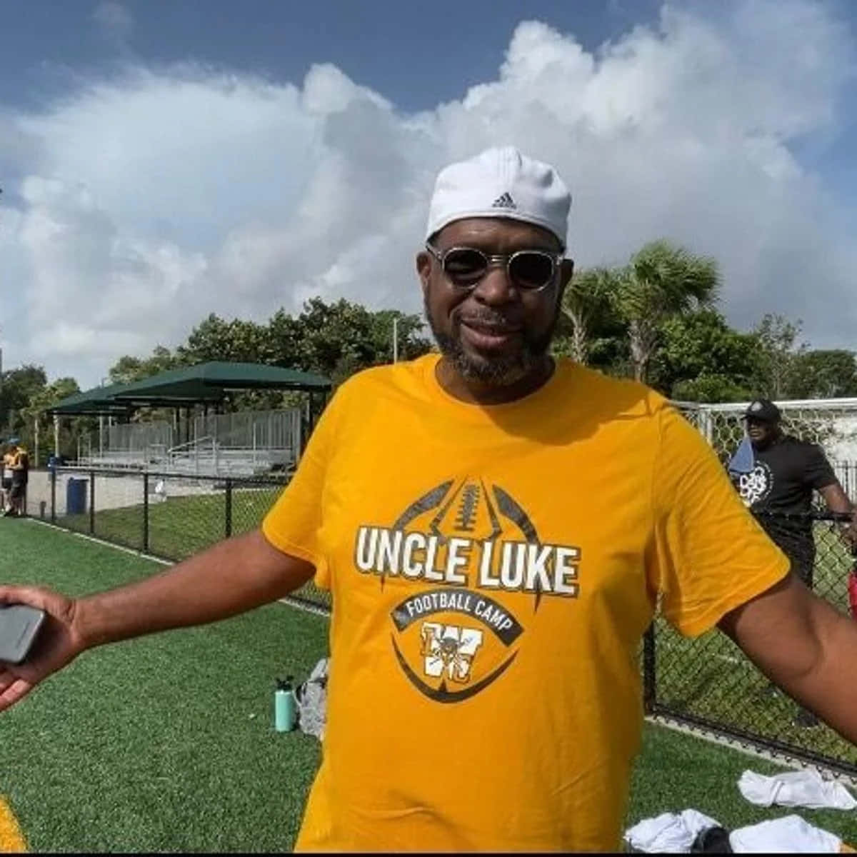  壁紙 — 一個微笑的男人穿著黃色「Uncle Luke Football Camp」襯衫和白色帽子，站在部分多雲的天空下的綠色草地場地上。