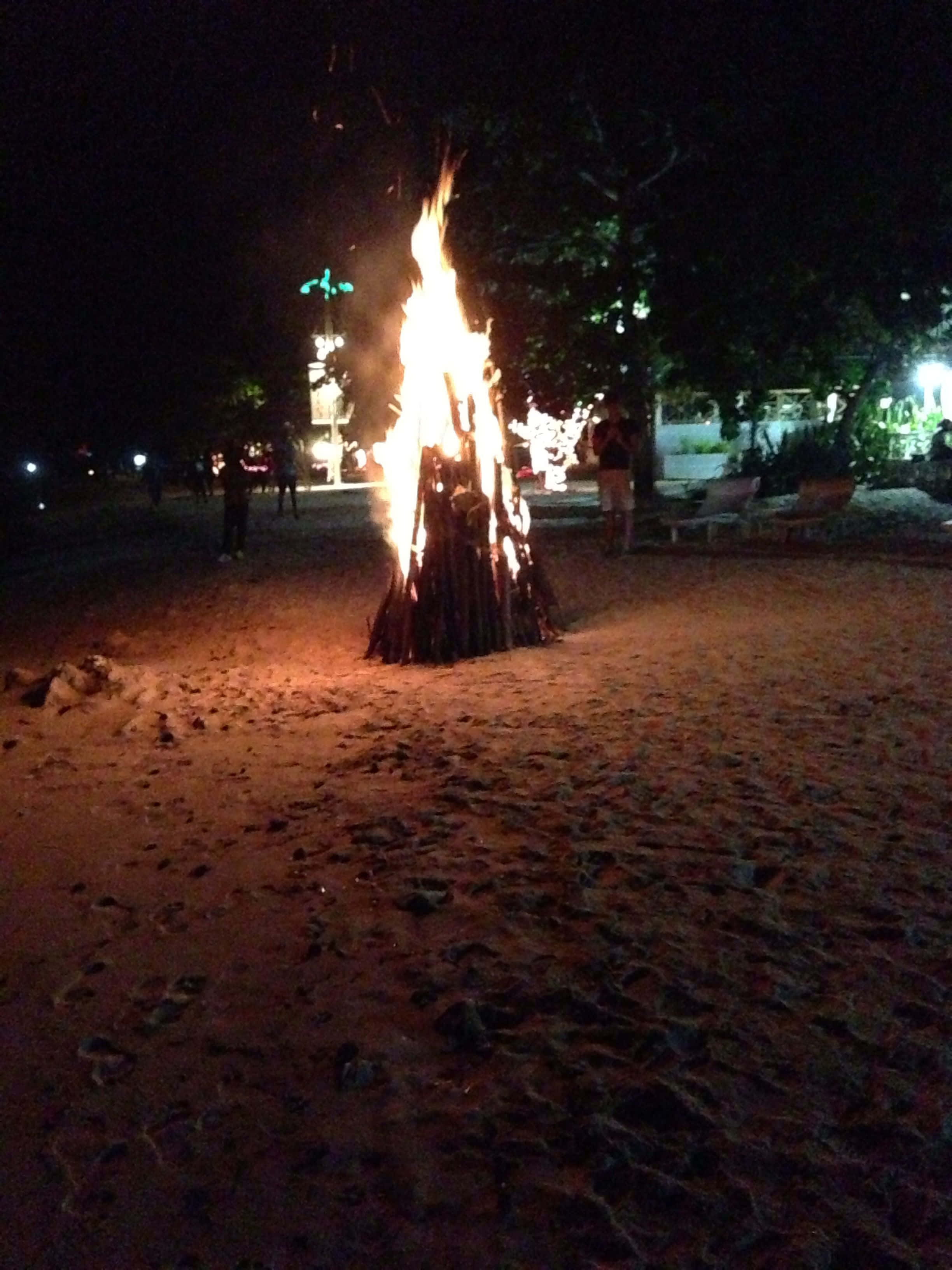 Beach Campfire 桌布 壁紙 — 一個熊熊燃燒的營火在夜晚的沙灘上明亮地燃燒，將溫暖的光芒投射在黑暗的環境和附近的剪影人物上。