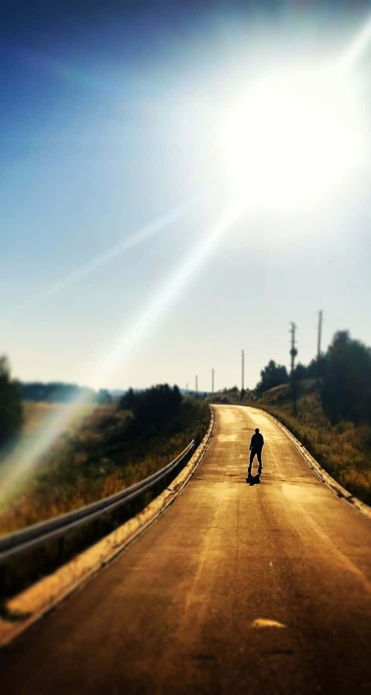  壁紙 — 一個孤獨的滑板手沿著陽光普照的鄉村道路滑行，明亮的光線穿透清澈的藍天。