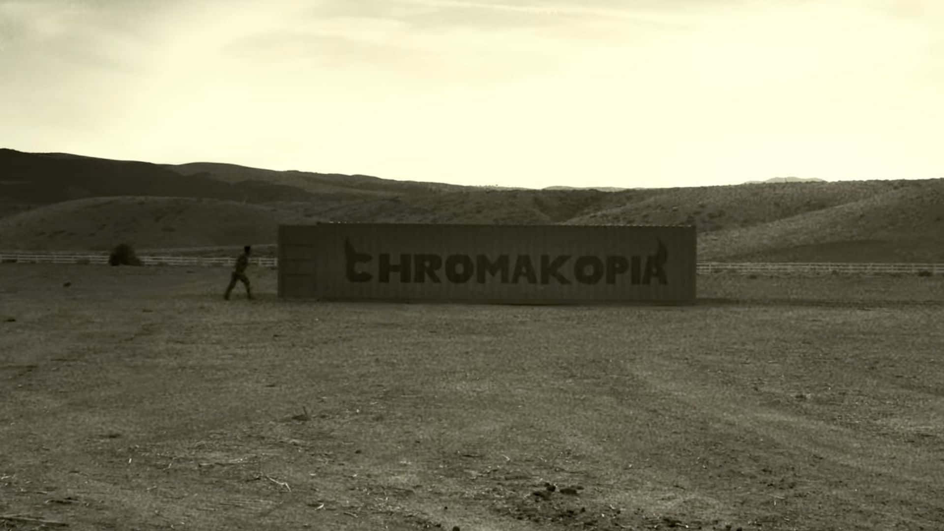  壁紙 — 一個孤獨的身影走向一個大型混凝土標誌，上面寫著 CHROMAKOPIA，背景是荒蕪的沙漠景觀，天空朦朧。