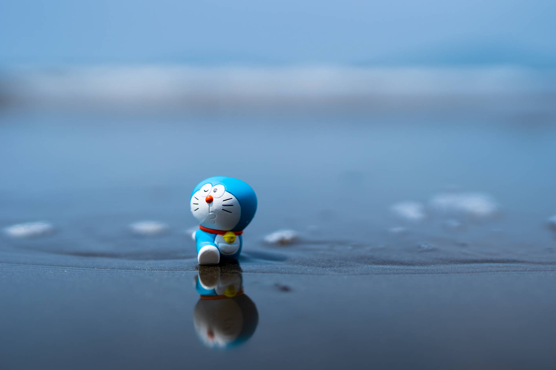 藍色多啦A夢公仔坐在濕沙灘上，帶有海洋倒影。