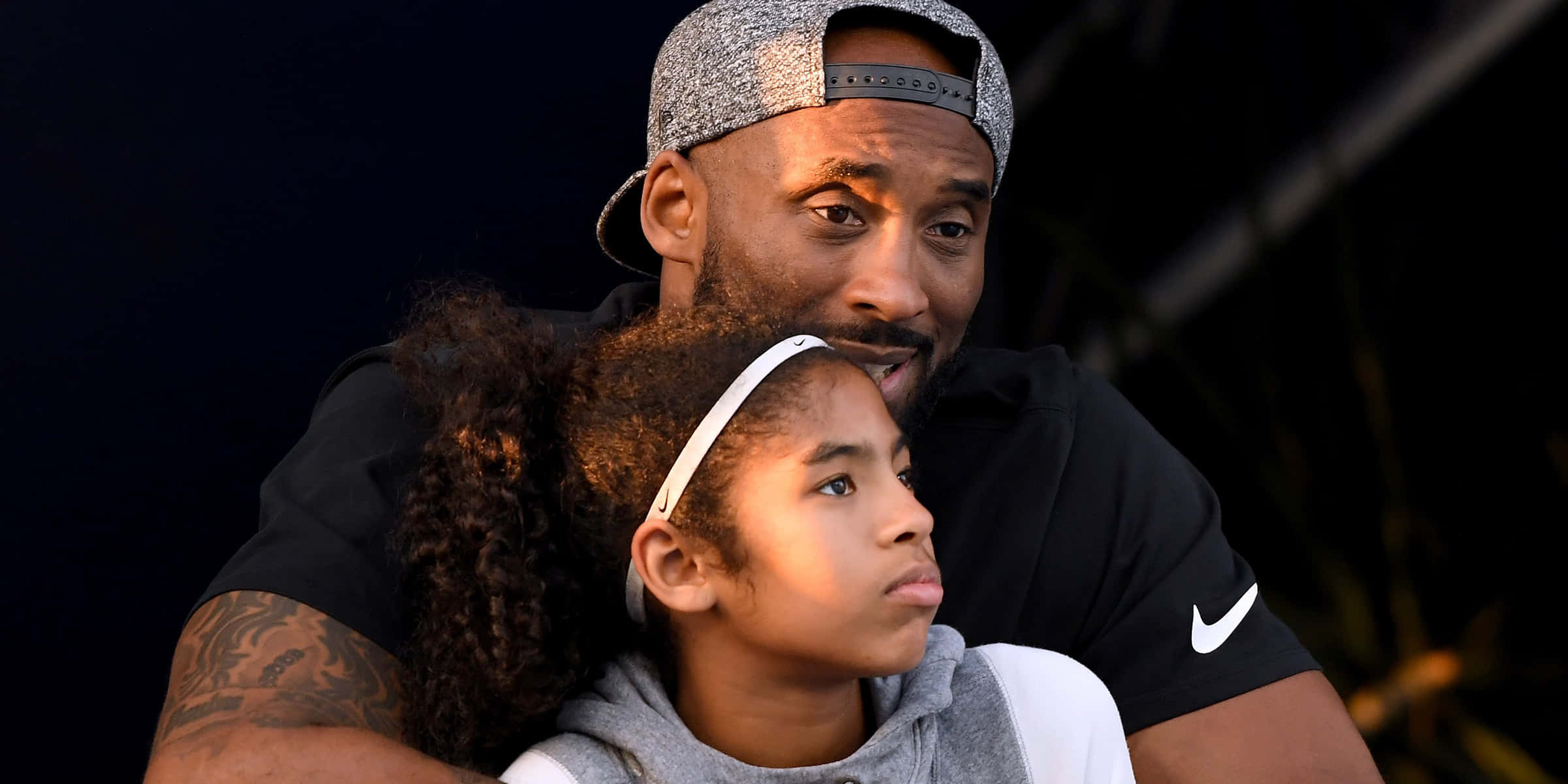 Kobe And Gigi 桌布 壁紙 — 一個有紋身手臂和戴反帽子的男人在深色背景前擁抱一個有捲髮和頭帶的小女孩。