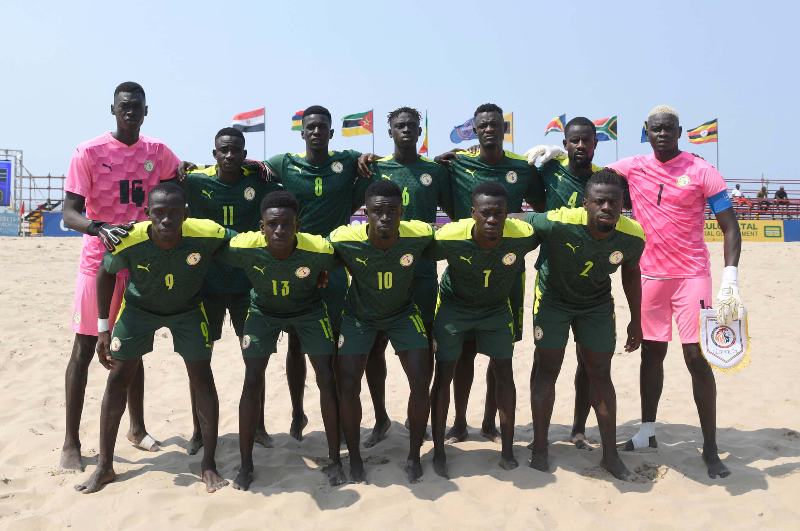 Beach Soccer 桌布 壁紙 — 一支身穿綠色和黃色球衣的沙灘足球隊在明亮的日光下自信地擺姿勢於沙地上，身後旗幟飄揚。