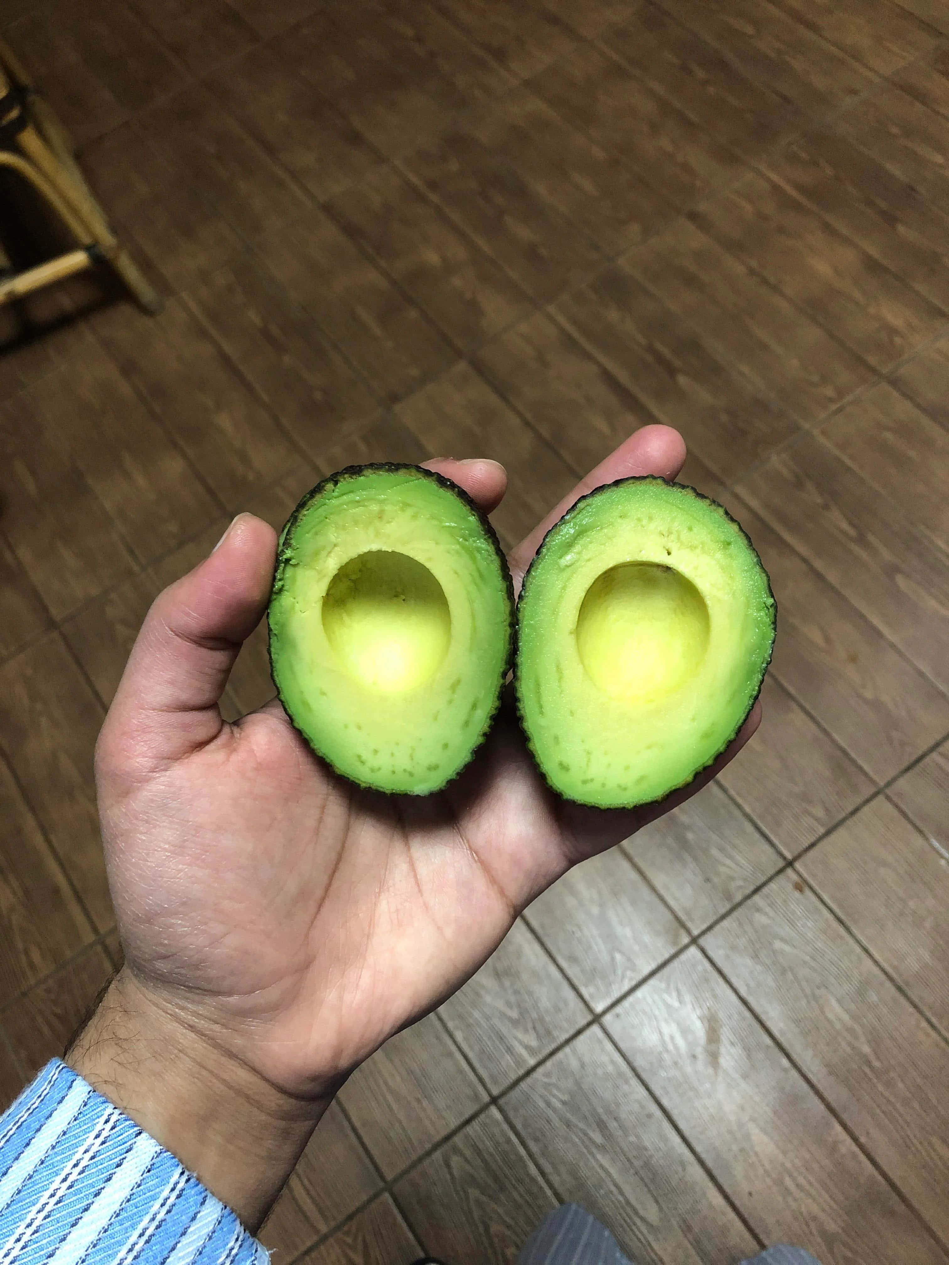 Avocado 桌布 壁紙 — 一隻手拿著兩個半邊成熟的牛油果，在木地板背景下露出奶油黃色的果肉。