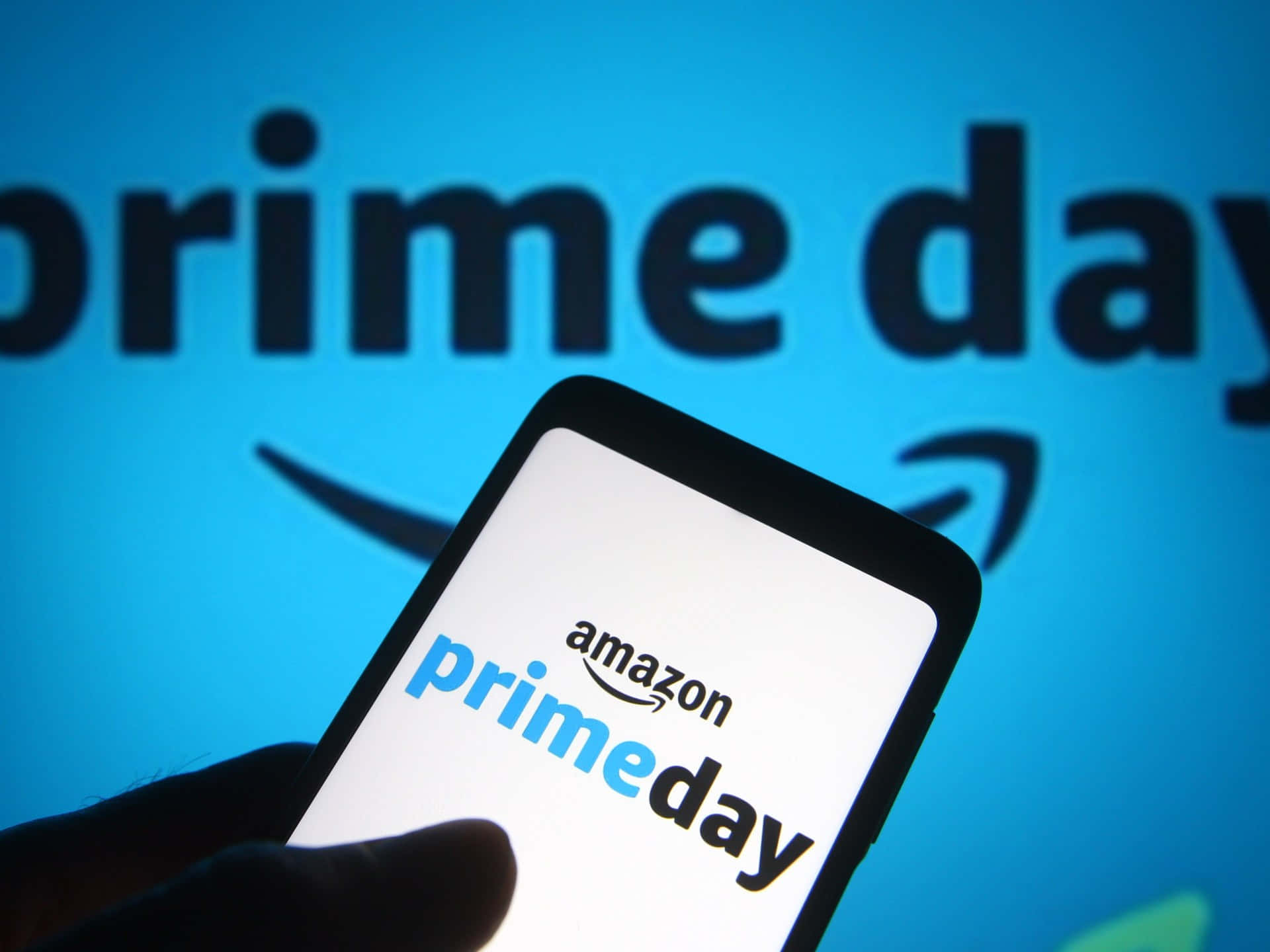  壁紙 — 一隻手拿著智能手機，屏幕上顯示Amazon Prime Day標誌，背景為藍色，帶有模糊的Prime Day文字和箭頭。