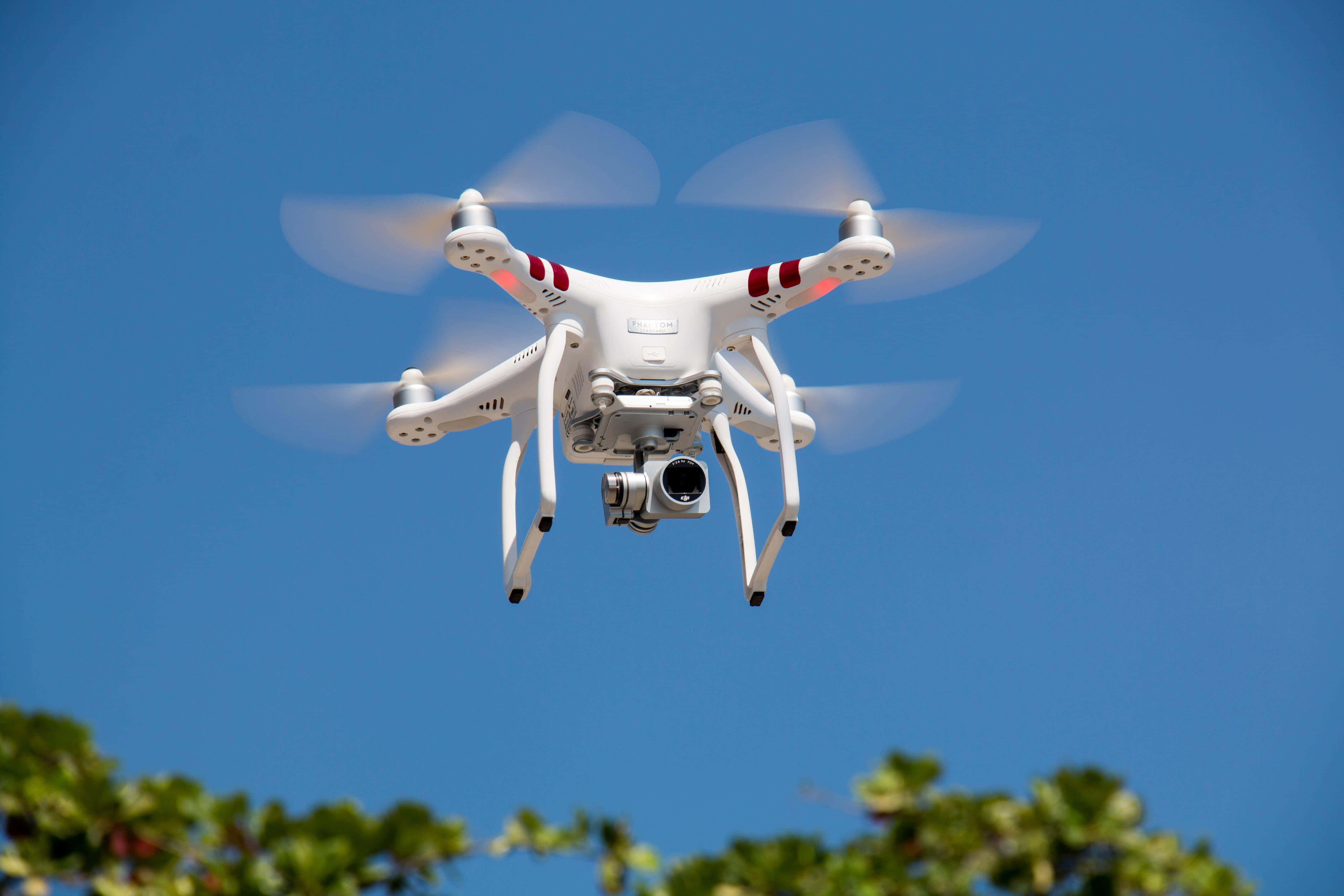 Drone Camera 桌布 壁紙 — 一架白色四軸無人機在清澈的藍天中盤旋，螺旋槳模糊，下方有綠色樹葉。