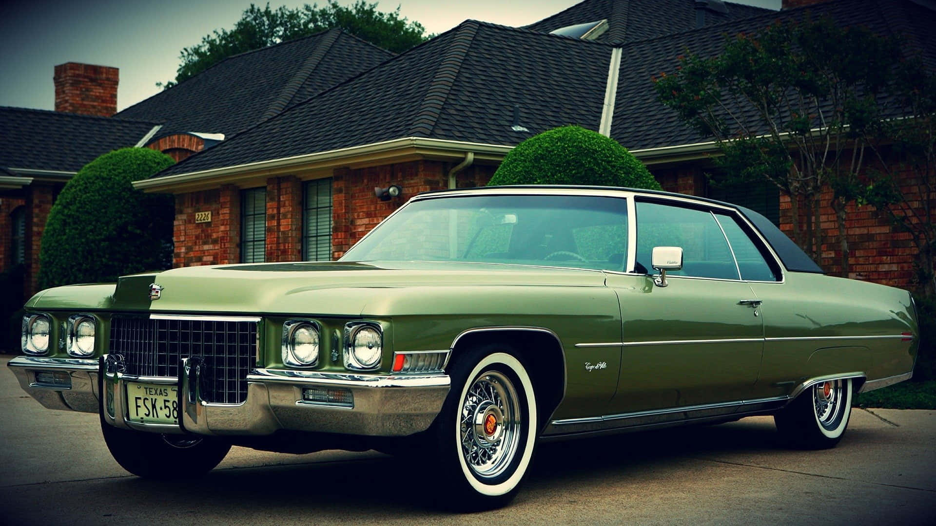 Cadillac Deville 桌布 壁紙 — 一架復古橄欖綠色的凱迪拉克 Coupe de Ville，配備白色牆壁輪胎，停在一間磚房前，周圍有修剪整齊的樹籬。