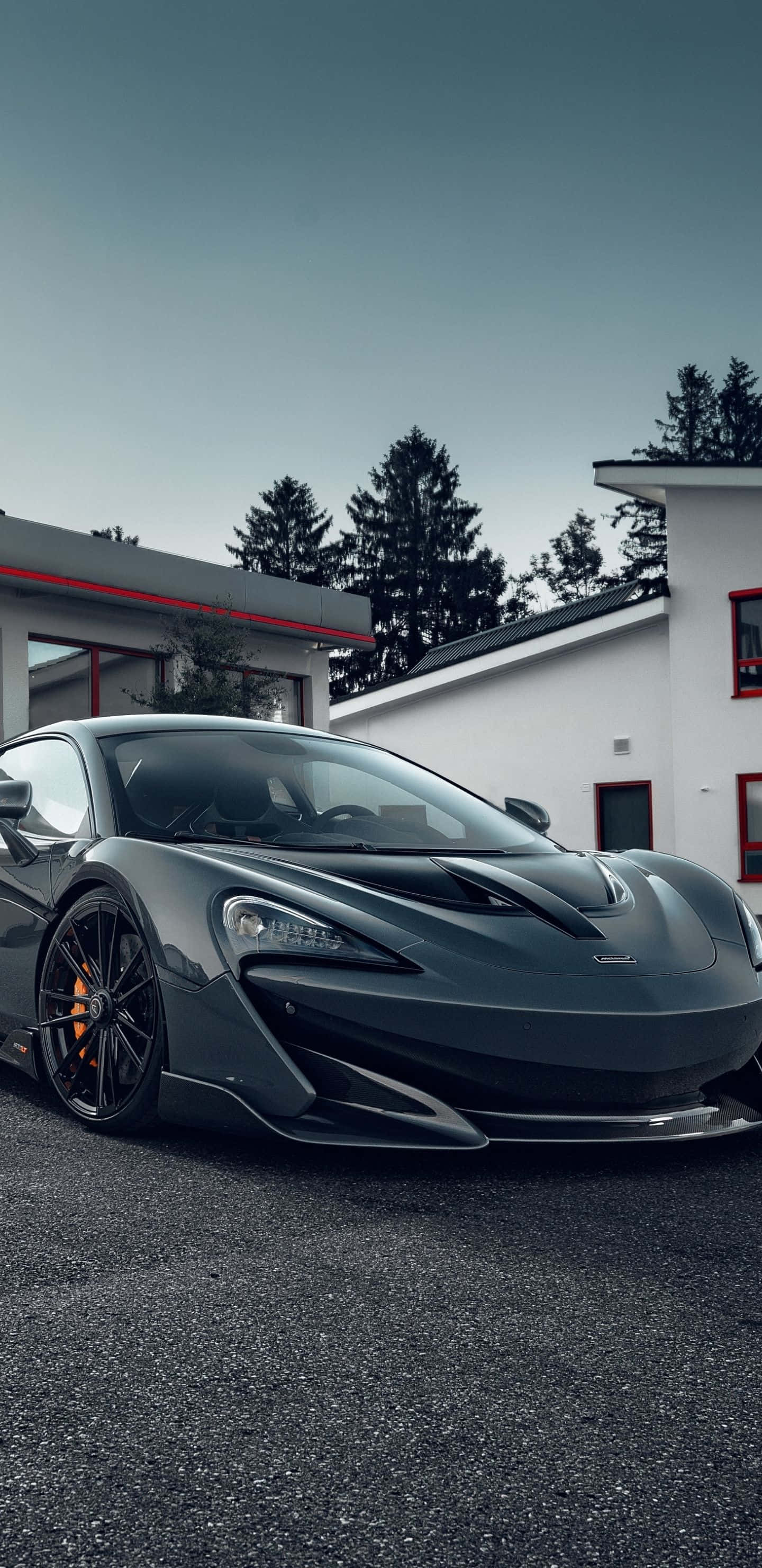 Mclaren 600lt 桌布 壁紙 — 一架時尚的深灰色McLaren跑車停泊在瀝青路上，前方是帶有紅色點綴的現代白色建築，天空陰沉多雲。