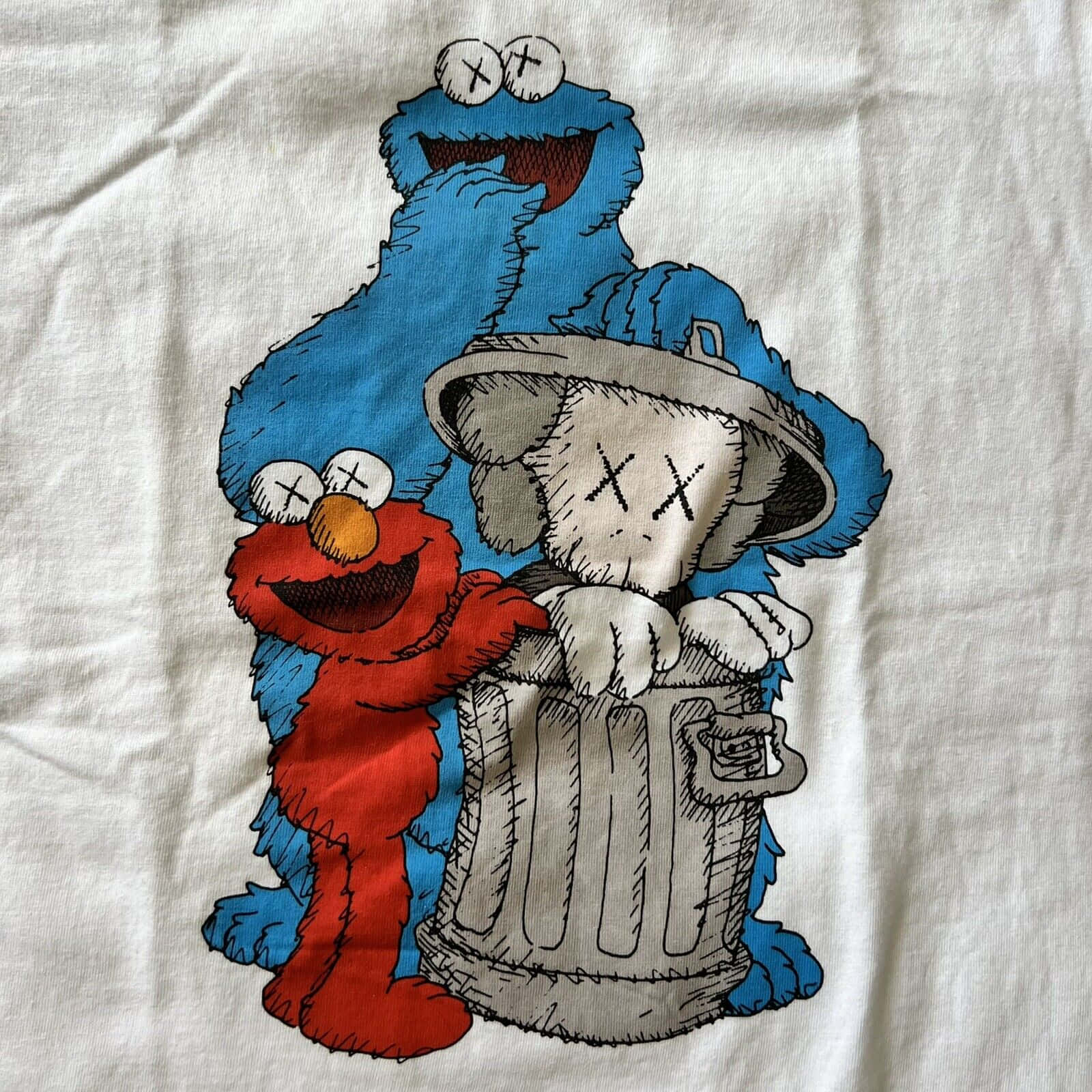 Kaws Sesame Street 桌布 壁紙 — 一件白色T恤上展示了一個圖案，圖案中Elmo和Cookie Monster擁抱一個帶有X形眼睛的KAWS風格垃圾桶角色。