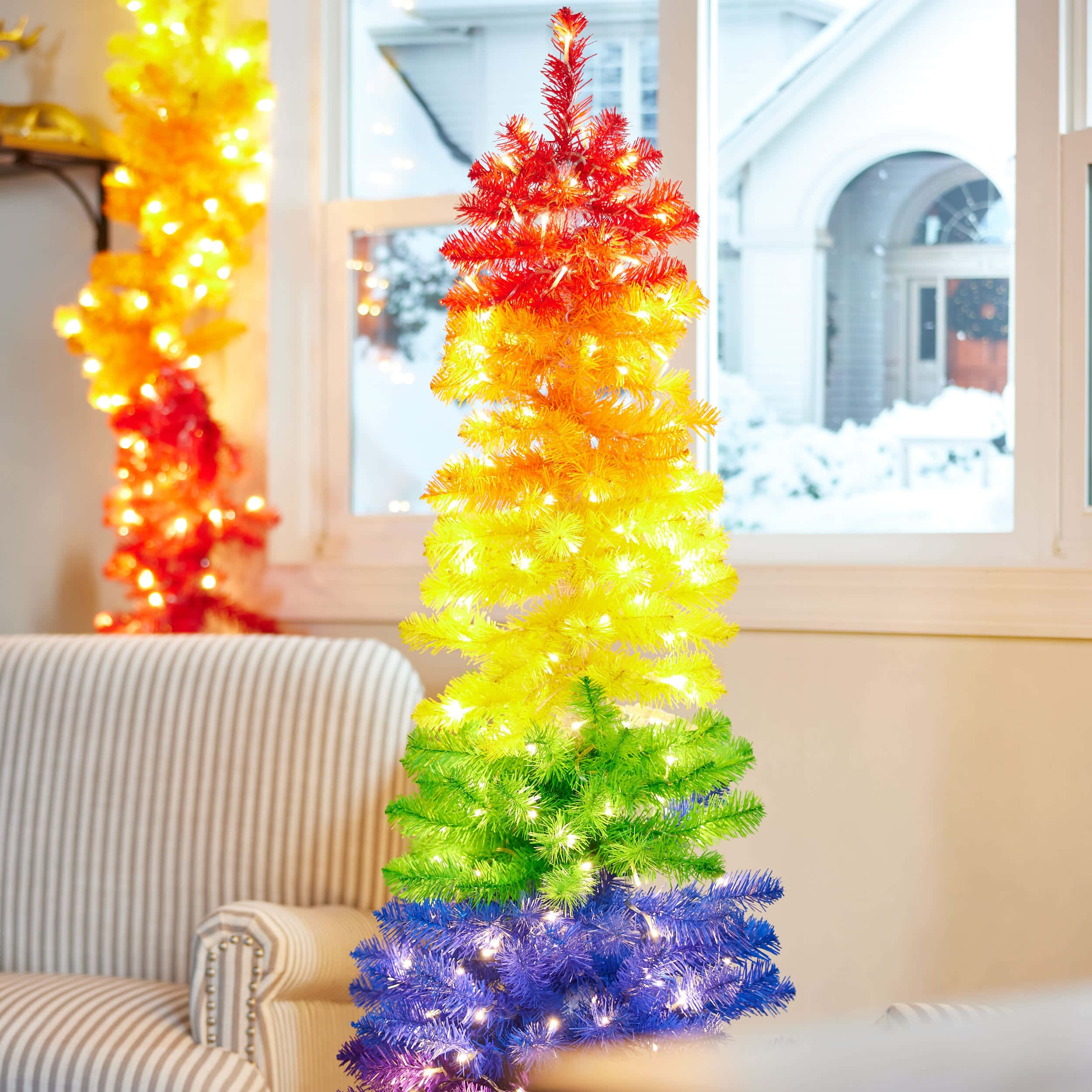 Christmas Rainbow 桌布 壁紙 — 一棵色彩繽紛的彩虹聖誕樹，帶有閃爍的燈光，放在室內靠近可看到雪景的窗邊。