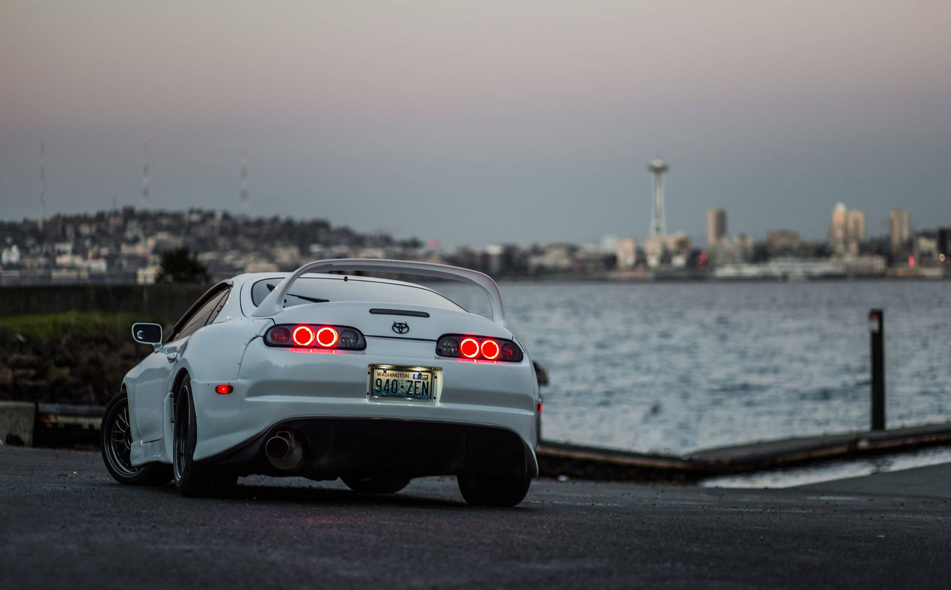 白色 Toyota Supra 停泊於海灣 壁紙 — 一輛白色豐田Supra，紅色尾燈發光，停在海濱，背景是黃昏時分的西雅圖天際線。