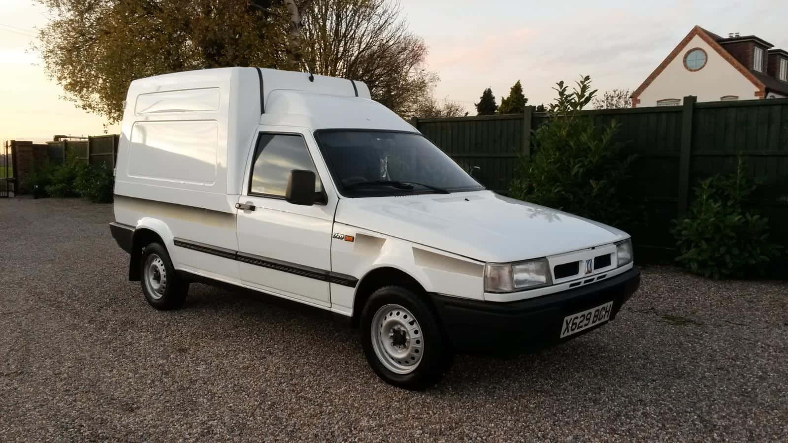 Fiat Fiorino 桌布 壁紙 — 一輛白色Fiat Ducato客貨車在黃昏時停泊在碎石上，背景有木柵欄和房屋。