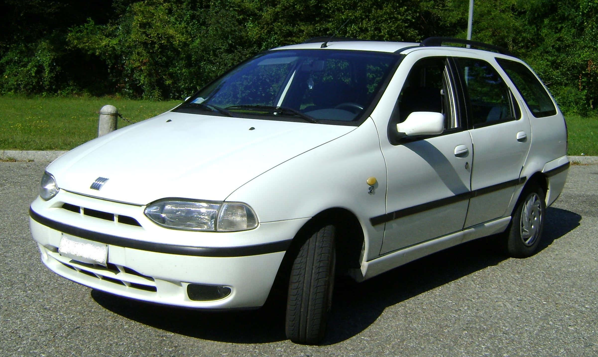 Fiat Palio 桌布 壁紙 — 一輛白色Fiat Stilo旅行車停在瀝青路上，背景有綠樹和草地，在明亮的日光下。