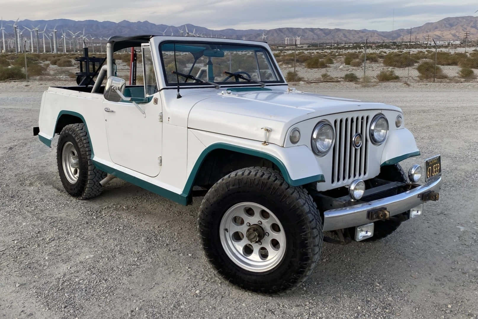 Jeepster 桌布 壁紙 — 一輛白色復古Jeep，帶有青綠色飾邊，停在礫石上，附近有風力發電機和乾燥山脈，天空多雲。