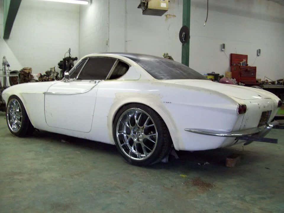 Volvo P1800 桌布 壁紙 — 一輛白色經典跑車配備鉻合金輪圈，停放在螢光燈下雜亂的車庫中。