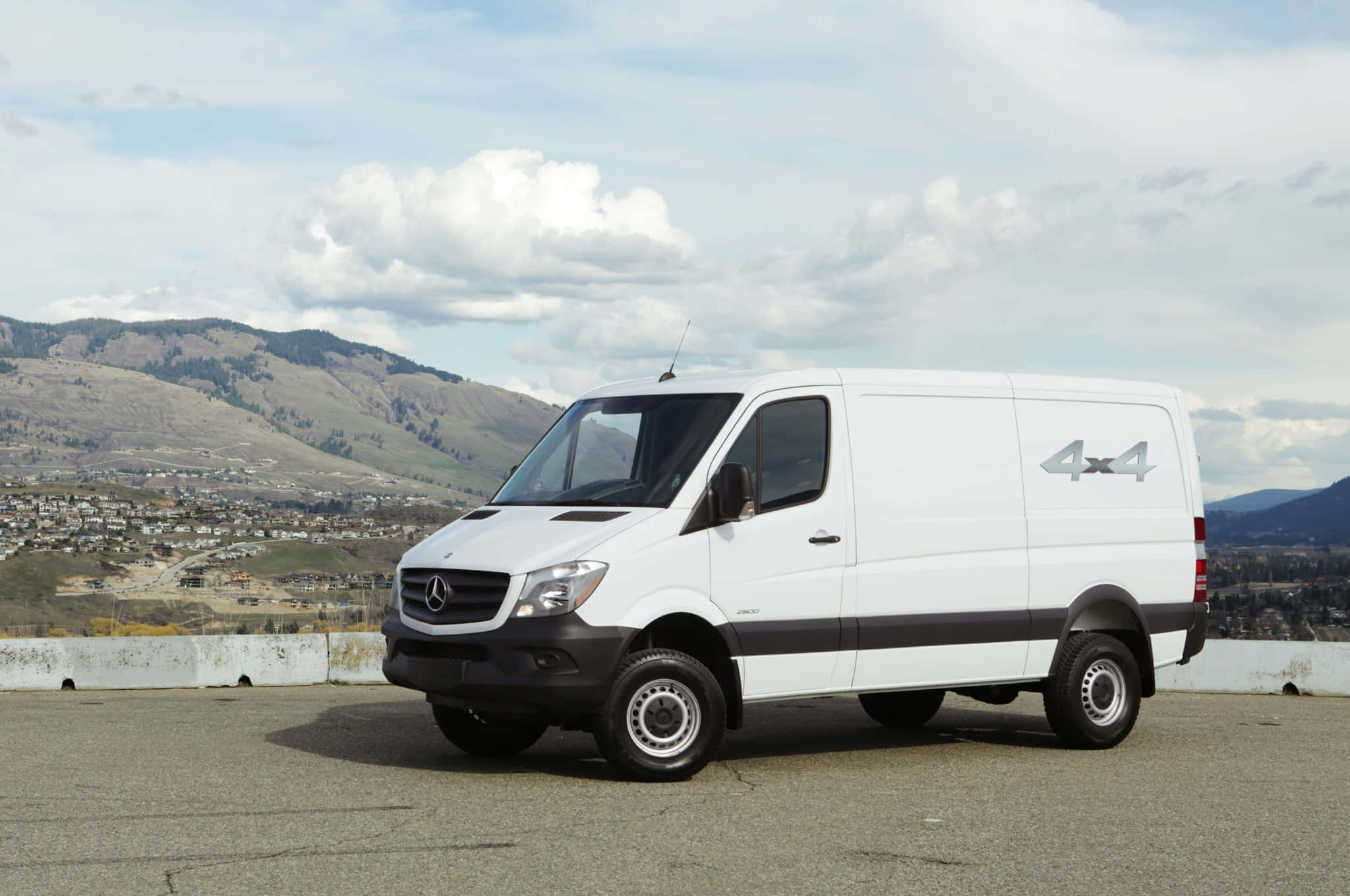 Mercedes Benz Sprinter 桌布 壁紙 — 一輛白色 Mercedes-Benz Sprinter 4x4 客貨車停泊在鋪砌的觀景點，下方是起伏的丘陵和城鎮，天空部分多雲。
