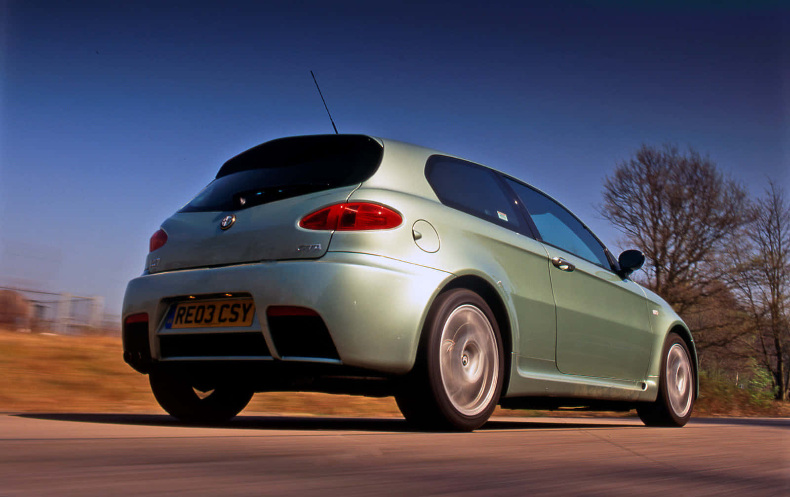 Alfa Romeo 147 桌布 壁紙 — 一輛薄荷綠色的愛快羅密歐147 GTA在晴朗的藍天下，以光禿的樹木為背景，沿著道路疾馳。