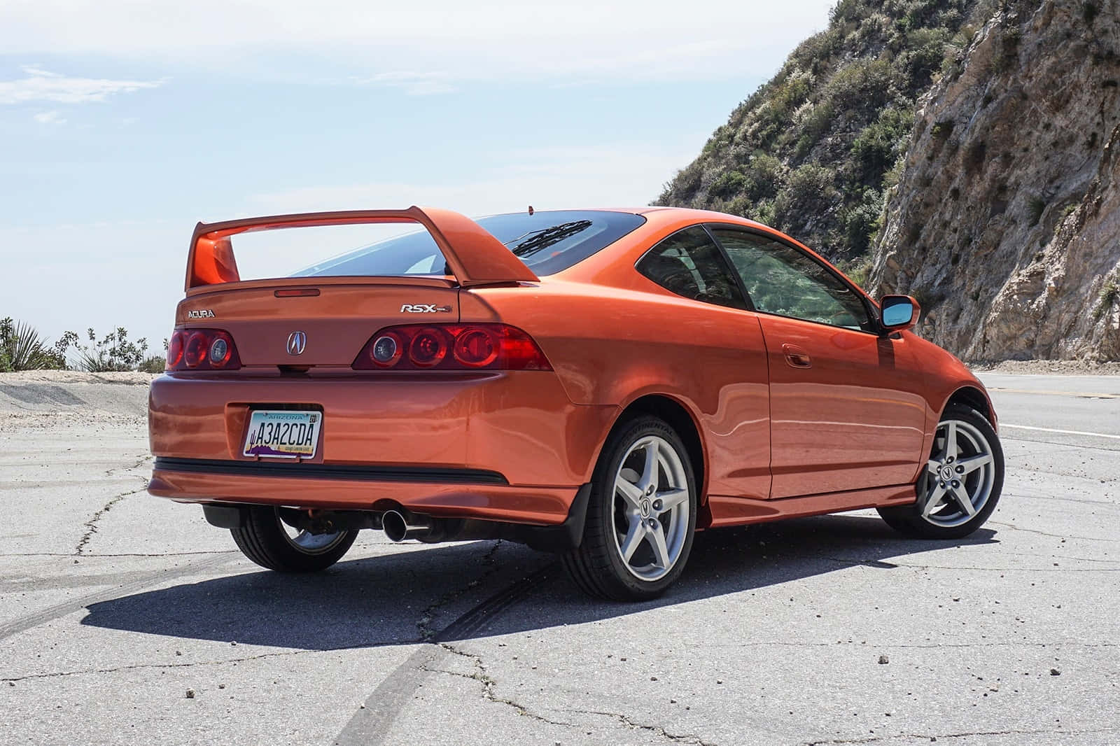 Acura Rsx 桌布 壁紙 — 一輛充滿活力的橙色Acura RSX Type-S雙門跑車停泊在裂開的瀝青路上，旁邊是岩石山丘，在清澈的藍天下。