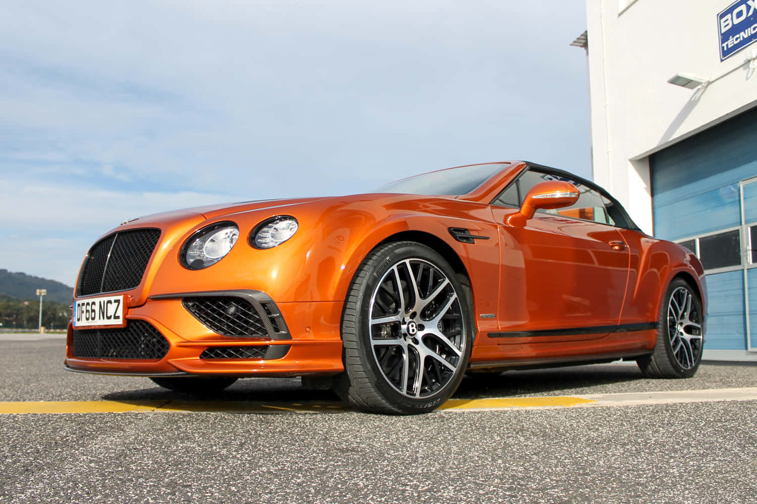 Bentley Continental Supersports 桌布 壁紙 — 一輛充滿活力的橙色賓利開篷車停泊在白色建築物旁邊的瀝青路面上，天空清澈湛藍。