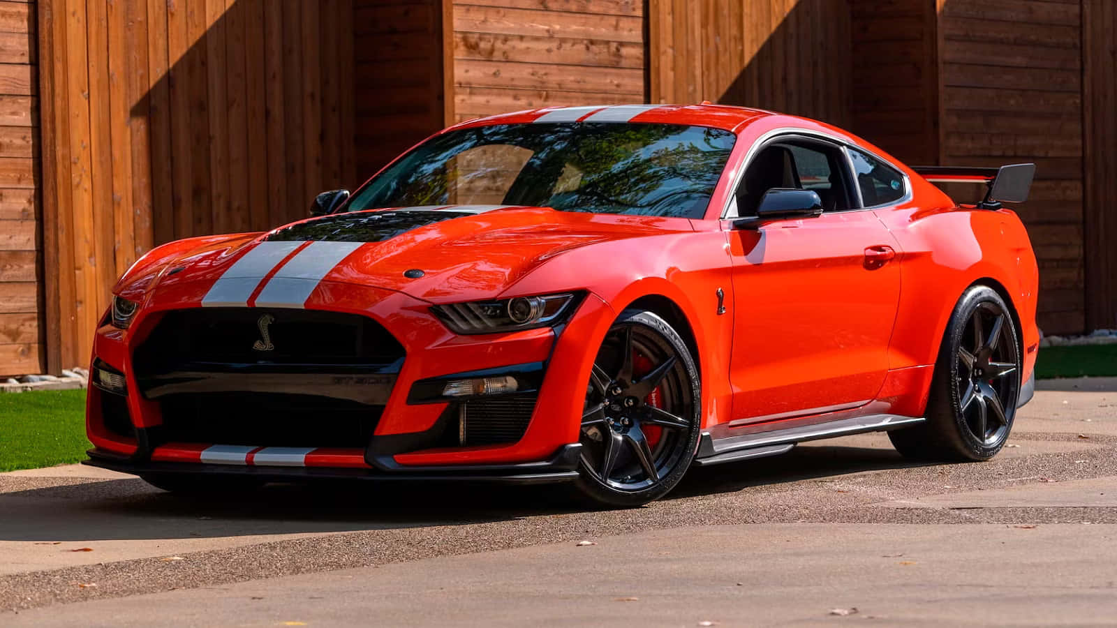  壁紙 — 一輛充滿活力的橙色福特野馬Shelby GT500，帶有白色賽車條紋，在陽光明媚的日子裡停在木車庫前。