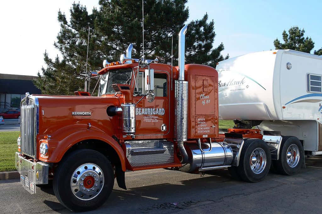 Kenworth C500 桌布 壁紙 — 一輛充滿活力的橙色Kenworth半掛卡車帶有鍍鉻裝飾，拖著一輛停在瀝青路面上的白色第五輪房車，背景是晴朗的藍天。