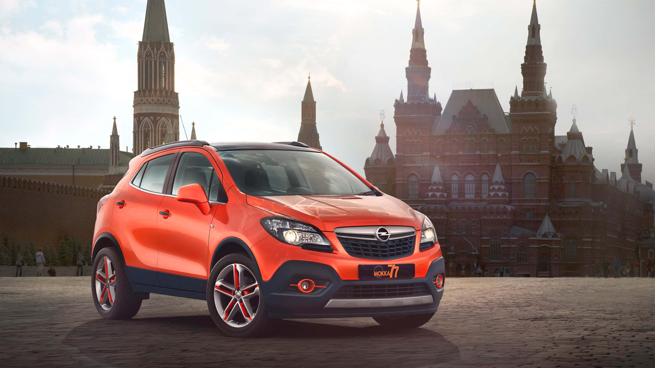 Opel Mokka 桌布 壁紙 — 一輛充滿活力的橙色Opel Mokka 7 SUV停在鵝卵石上，前方是歷史悠久的紅磚塔樓，天空柔和，光線明亮。