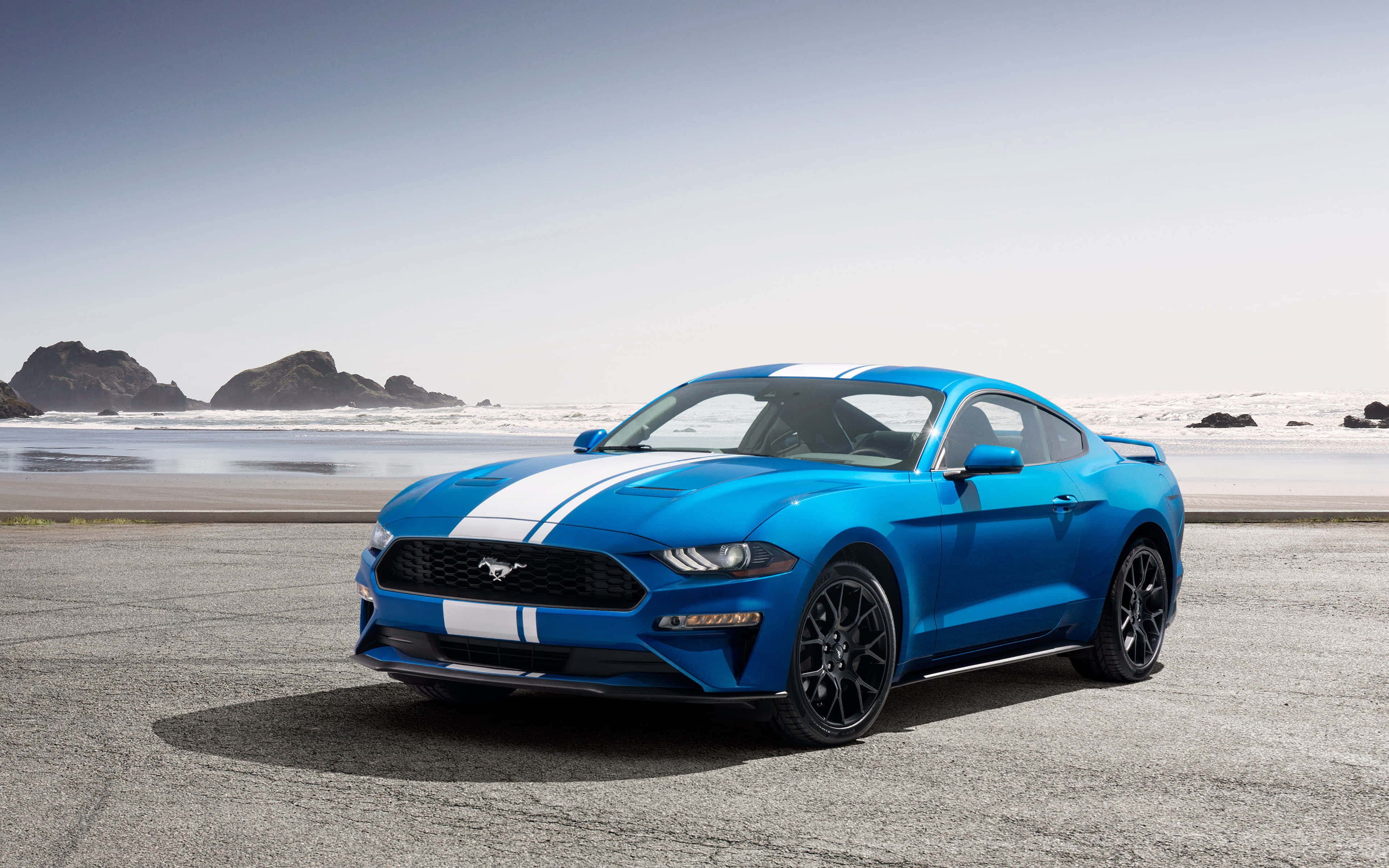 Ford Mustang Ecoboost 桌布 壁紙 — 一輛充滿活力的藍色福特野馬，帶有白色賽車條紋，停在沿海的碎石地上，背景是海浪和岩石岬角。