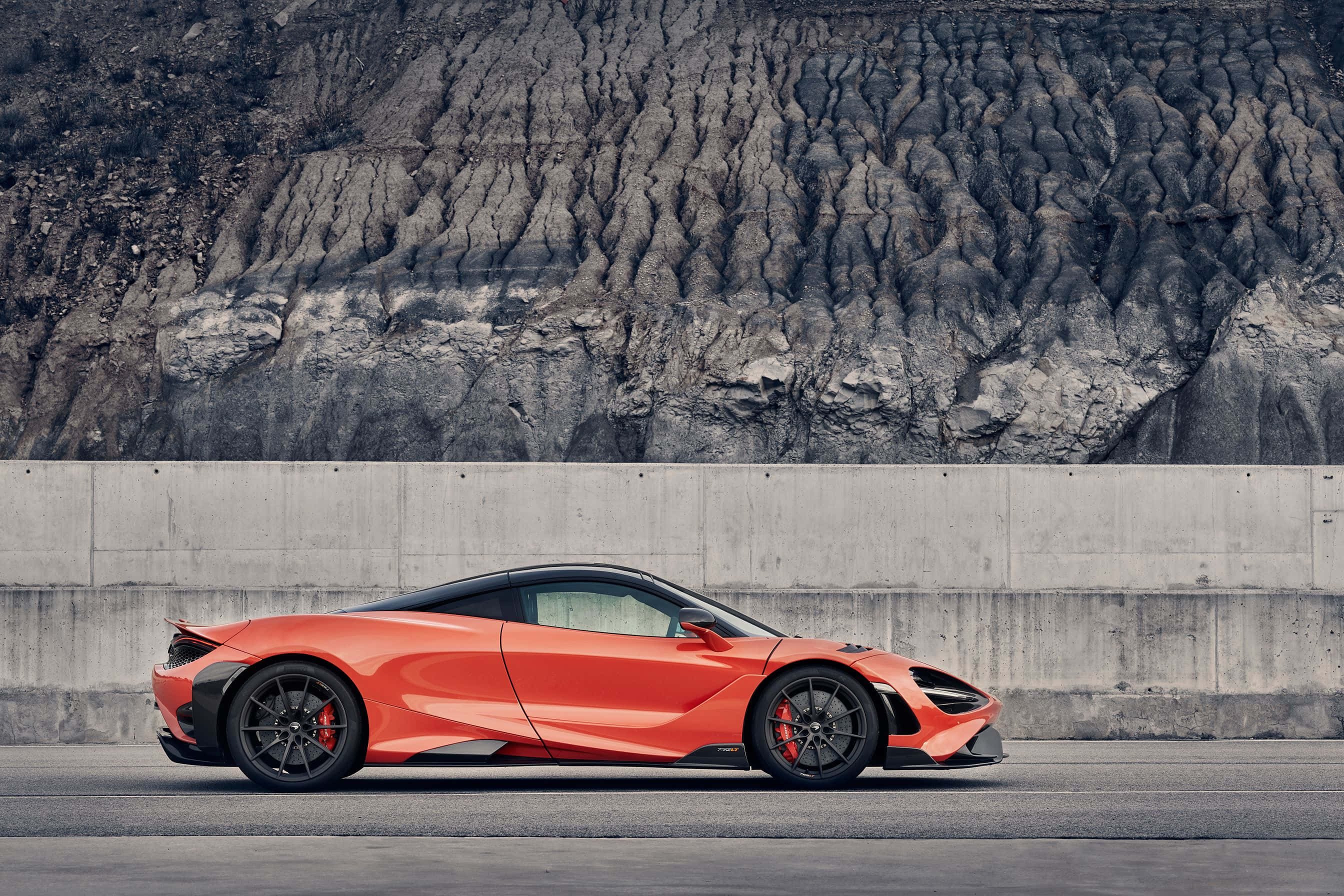 Mclaren 765lt 桌布 壁紙 — 一輛充滿活力的橙色McLaren跑車停在瀝青路面上，前方是崎嶇的灰色岩石懸崖。