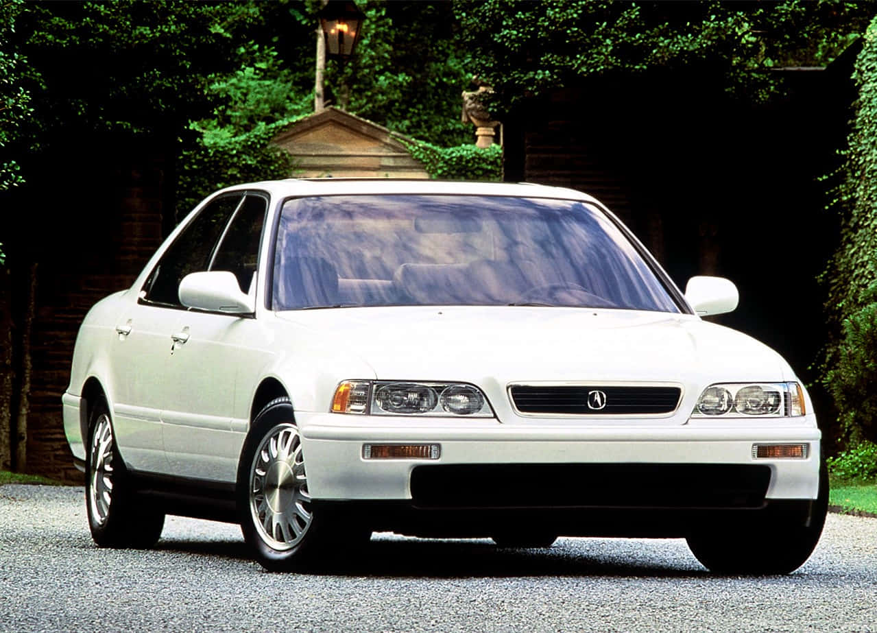 Acura Legend 桌布 壁紙 — 一輛純白色的Acura房車停泊在碎石車道上，周圍環繞著茂密的綠植和一座石屋。