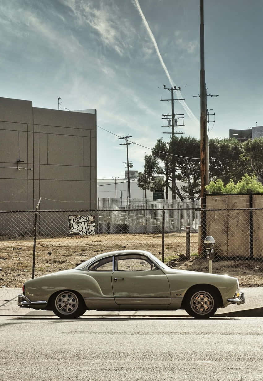  壁紙 — 一輛復古橄欖綠色的Karmann Ghia雙門跑車停泊在陽光普照的城市街道上，旁邊有鐵絲網圍欄和電線。