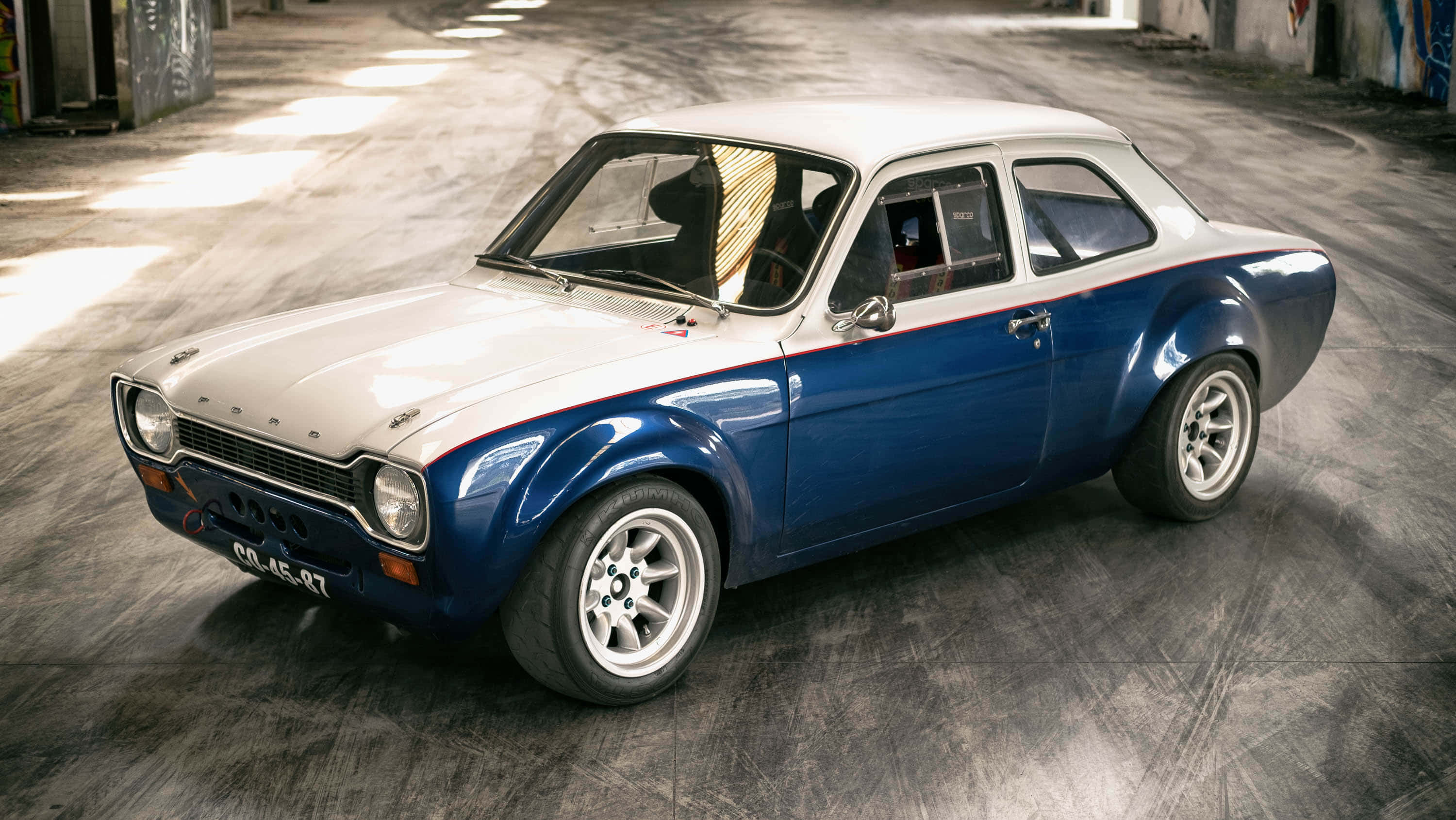 Ford Escort 桌布 壁紙 — 一輛復古藍白福特Escort拉力賽車停放在粗糙的混凝土車庫中，陽光照射進來。