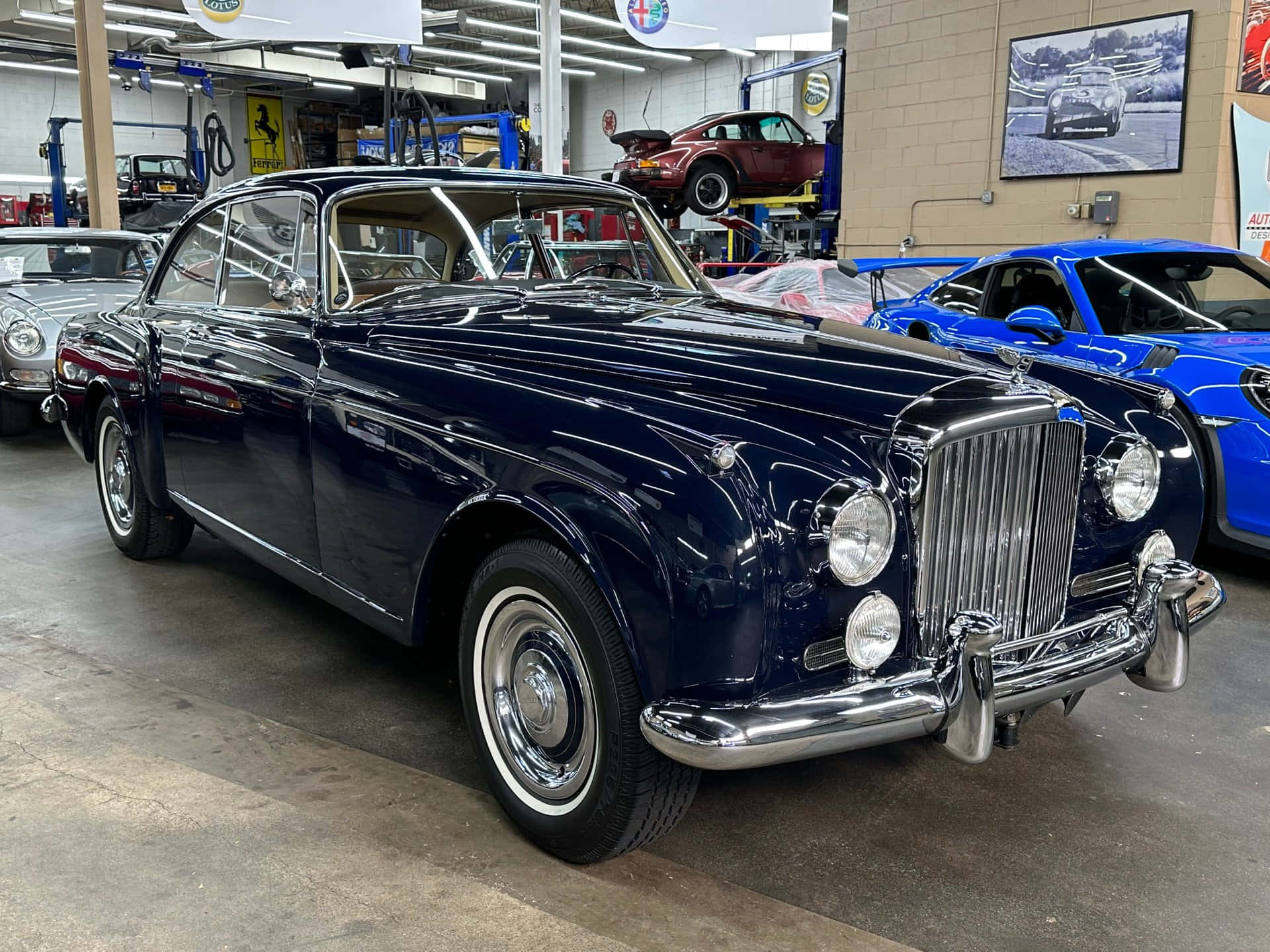 Bentley S2 桌布 壁紙 — 一輛光澤的海軍藍經典汽車在明亮的車庫燈光下閃閃發光，周圍環繞著其他復古車輛。