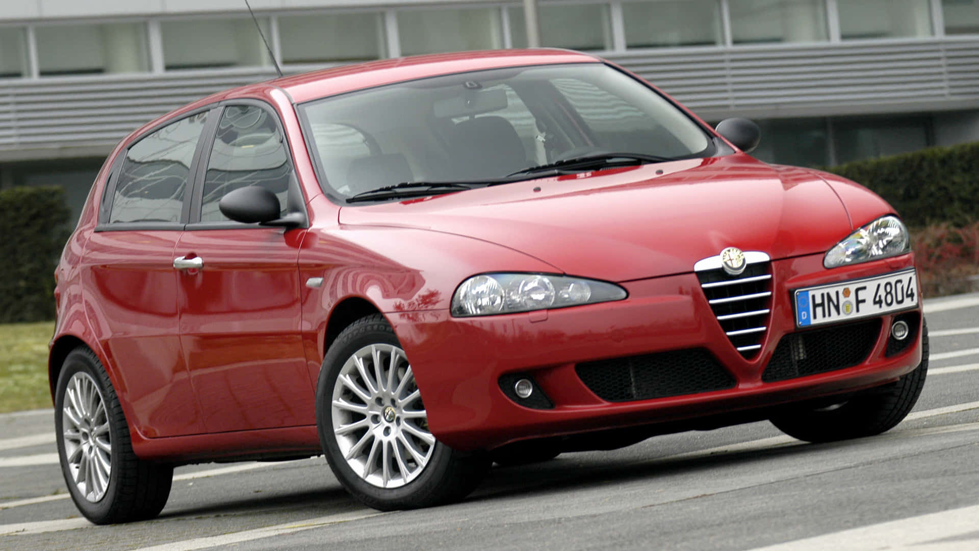 Alfa Romeo 147 桌布 壁紙 — 一輛光澤的紅色愛快羅密歐掀背車停在瀝青路上，背景有現代建築和綠化。