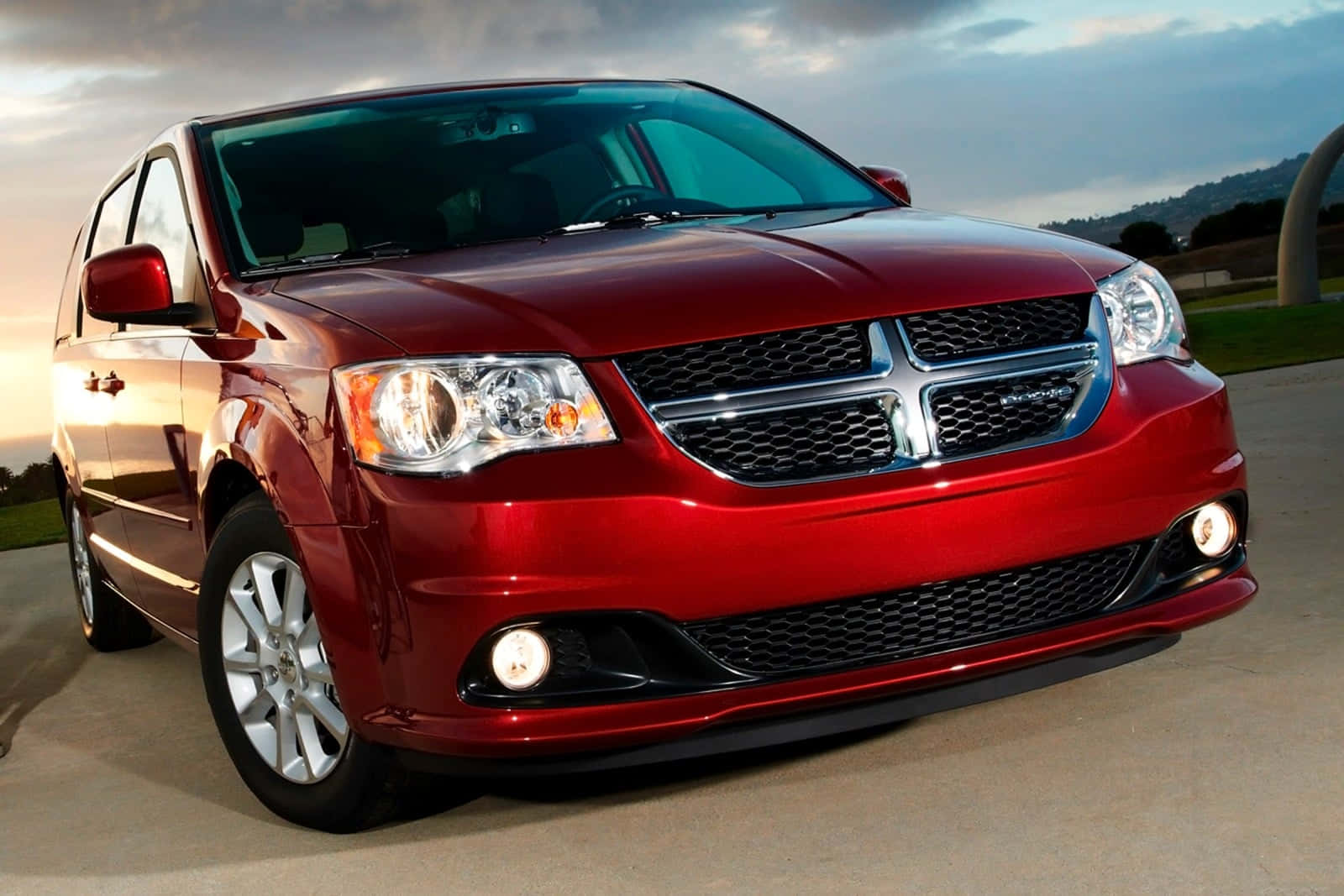  壁紙 — 一輛光澤的紅色道奇Grand Caravan小型貨車在金色日落光線下閃耀，背景是丘陵和鋪砌的車道。
