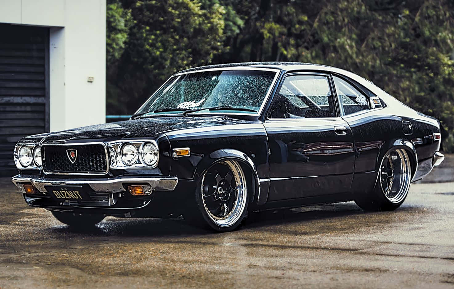Mazda Rx-3 桌布 壁紙 — 一輛光澤黑色經典肌肉車配備定制輪圈，在車庫旁的濕混凝土表面上於柔和燈光下閃閃發光。