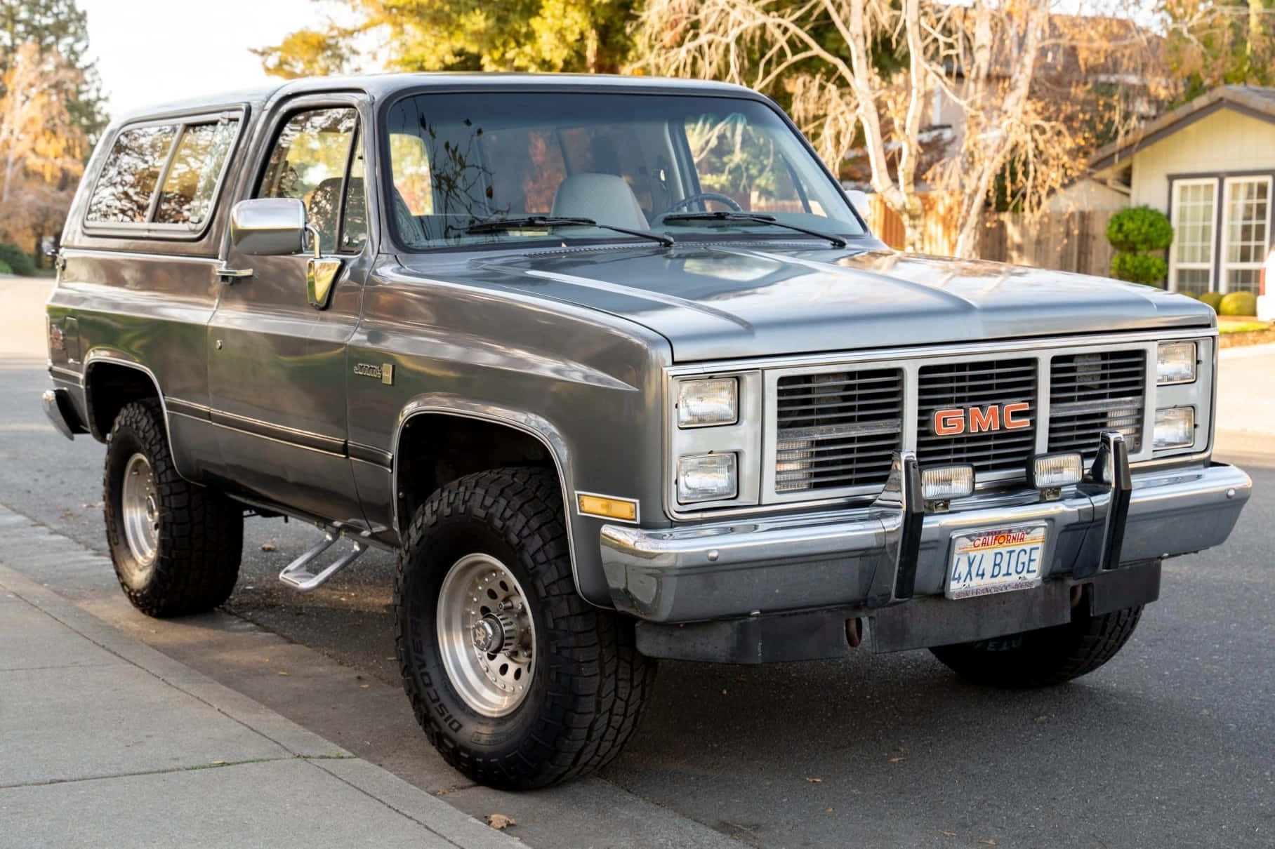 Gmc Jimmy 桌布 壁紙 — 一輛灰色的1980年代GMC Suburban SUV停泊在郊區街道上，背景有樹木和房屋。