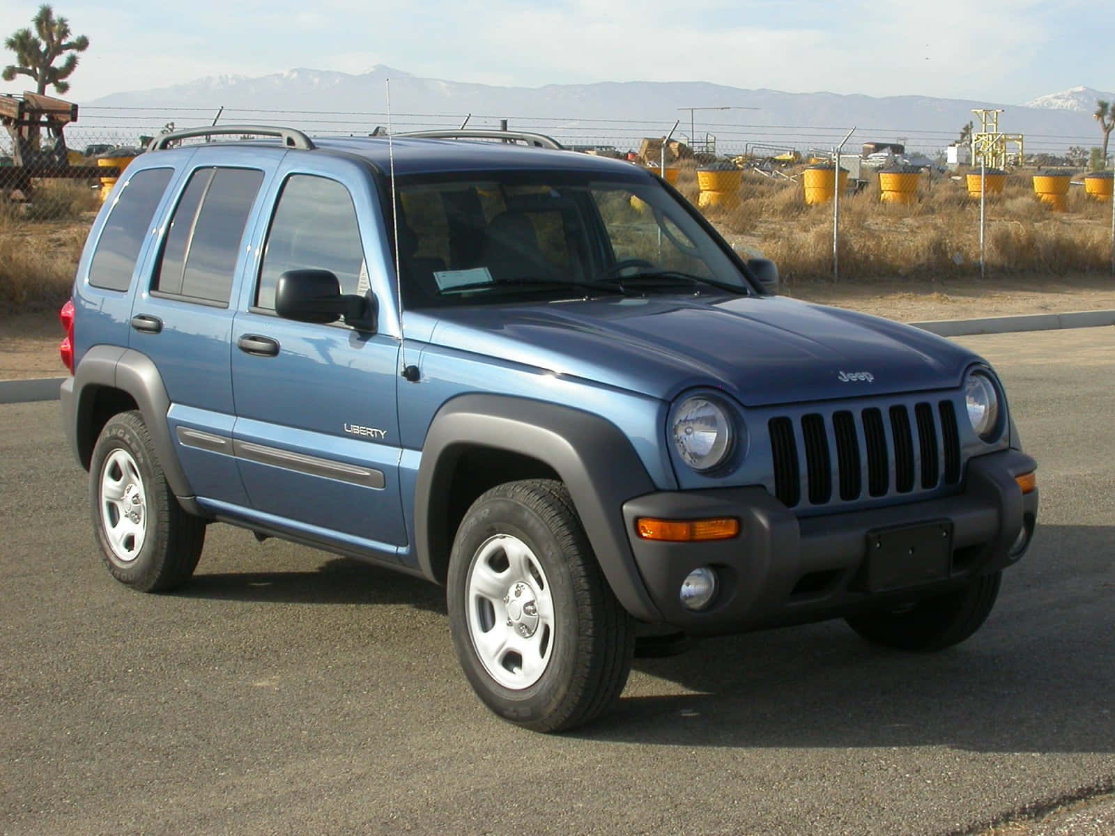Jeep Liberty 桌布 壁紙 — 一輛藍色Jeep Liberty SUV停泊在瀝青路上，背景有山脈和黃色桶，天空晴朗。