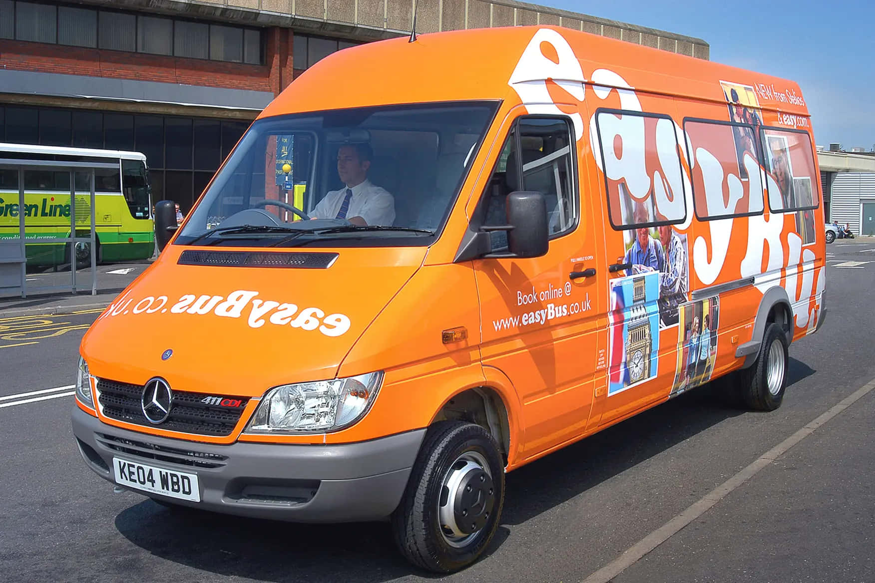 Mercedes Benz Sprinter 桌布 壁紙 — 一輛亮橙色的EasyBus Mercedes Sprinter客貨車停泊在巴士站，司機在車內，背景可見一輛綠色巴士。