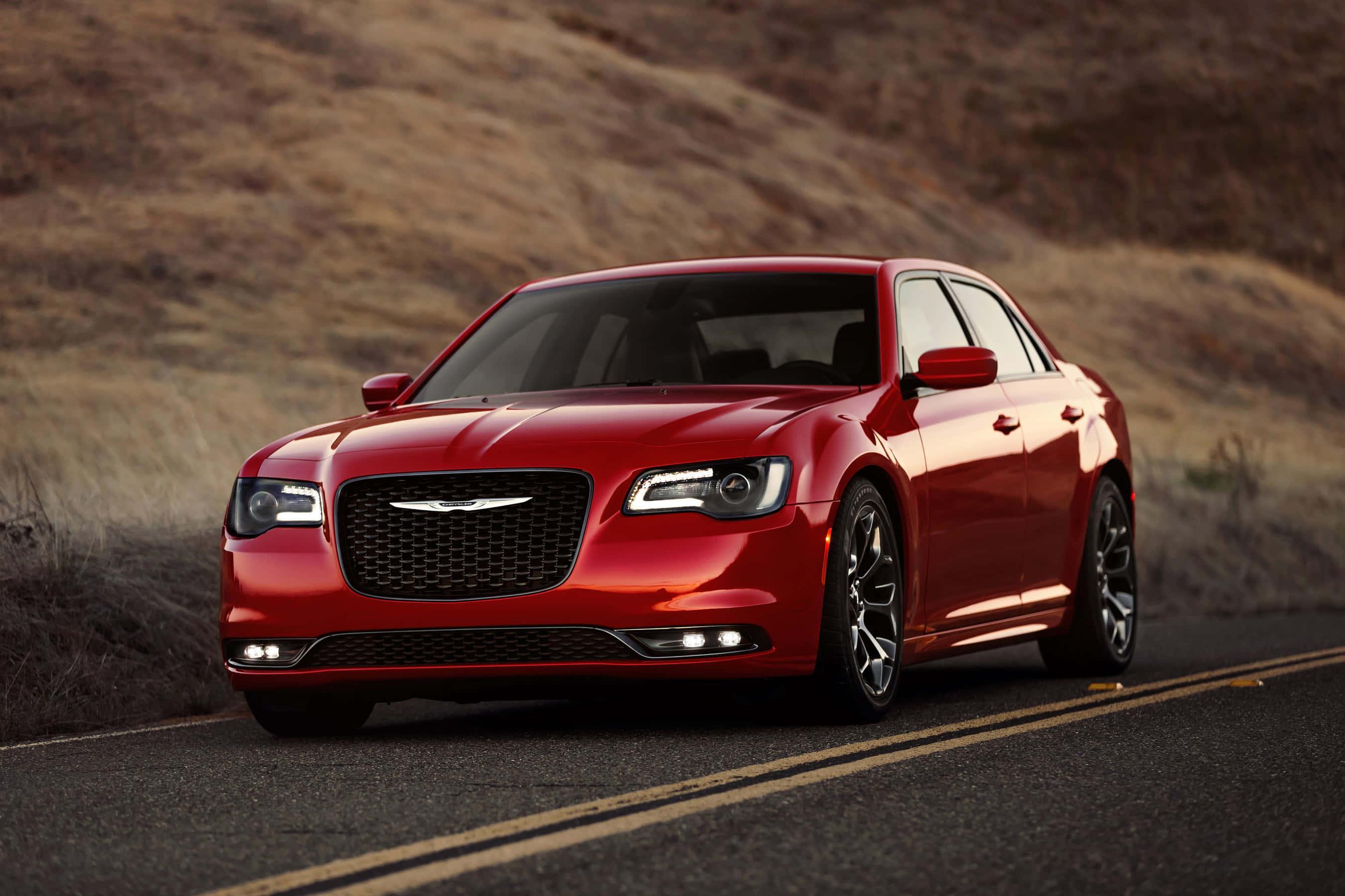 Chrysler 300 桌布 壁紙 — 一輛亮紅色的克萊斯勒轎車沿著蜿蜒的瀝青路行駛，穿過乾燥的金色山丘，沐浴在溫暖的陽光下。