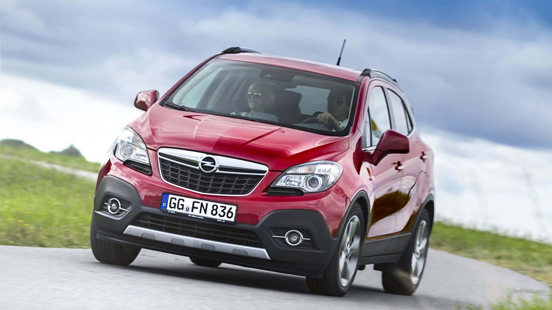 Opel Mokka 桌布 壁紙 — 一輛亮紅色的Opel Mokka SUV沿著蜿蜒的道路行駛，背景是綠色山丘和多雲的天空。