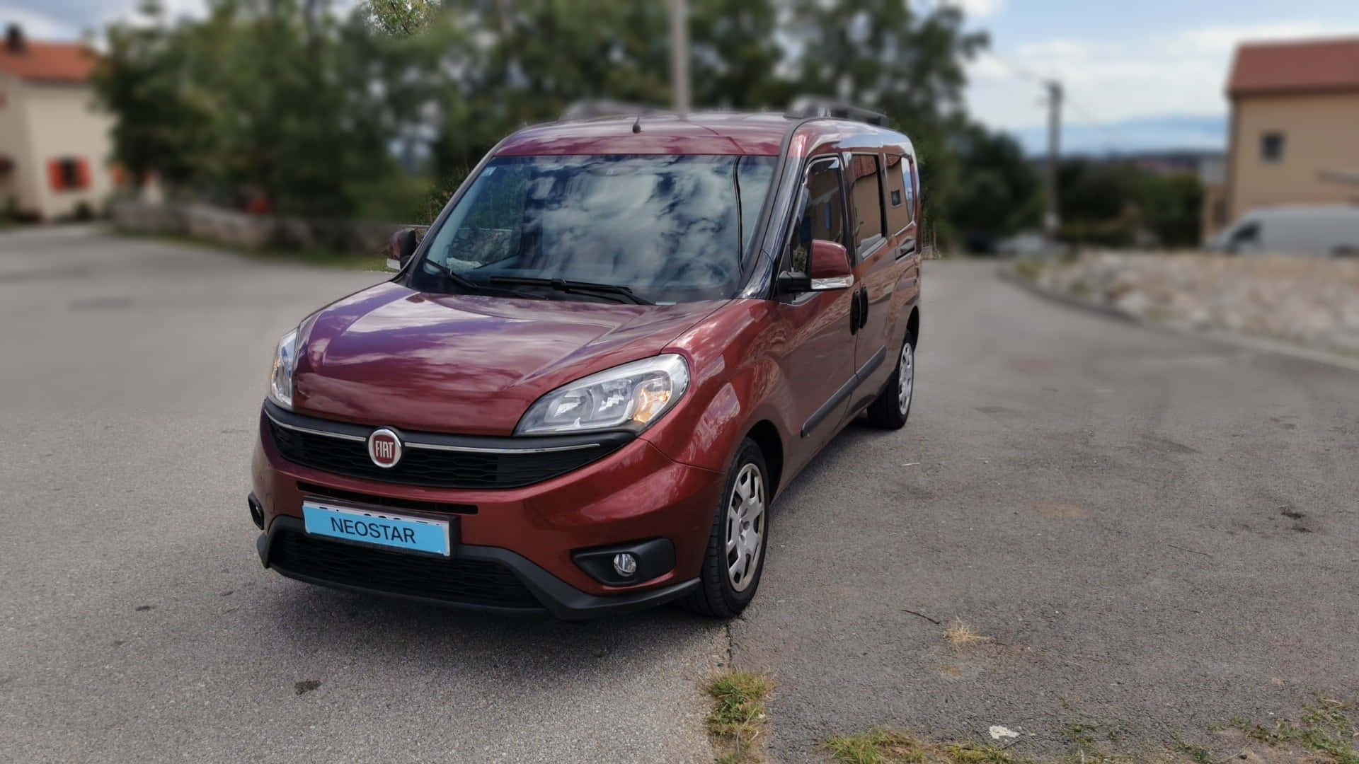  壁紙 — 一輛亮紅色的 Fiat Neostar 客貨車停泊在寧靜的瀝青路上，背景有樹木和房屋。