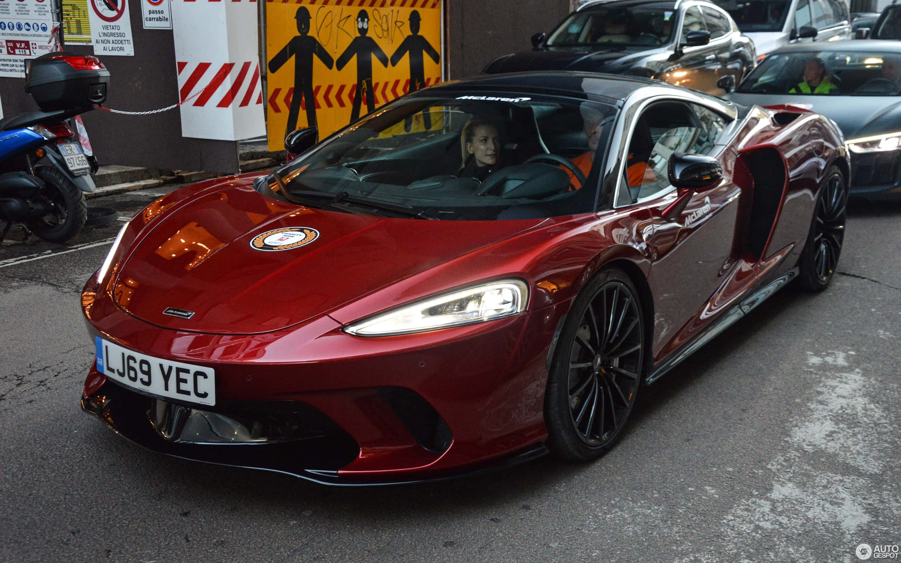 Mclaren Gt 桌布 壁紙 — 一輛亮紅色McLaren跑車打開車門，停在繁忙的城市街道上，車內有一位女士。