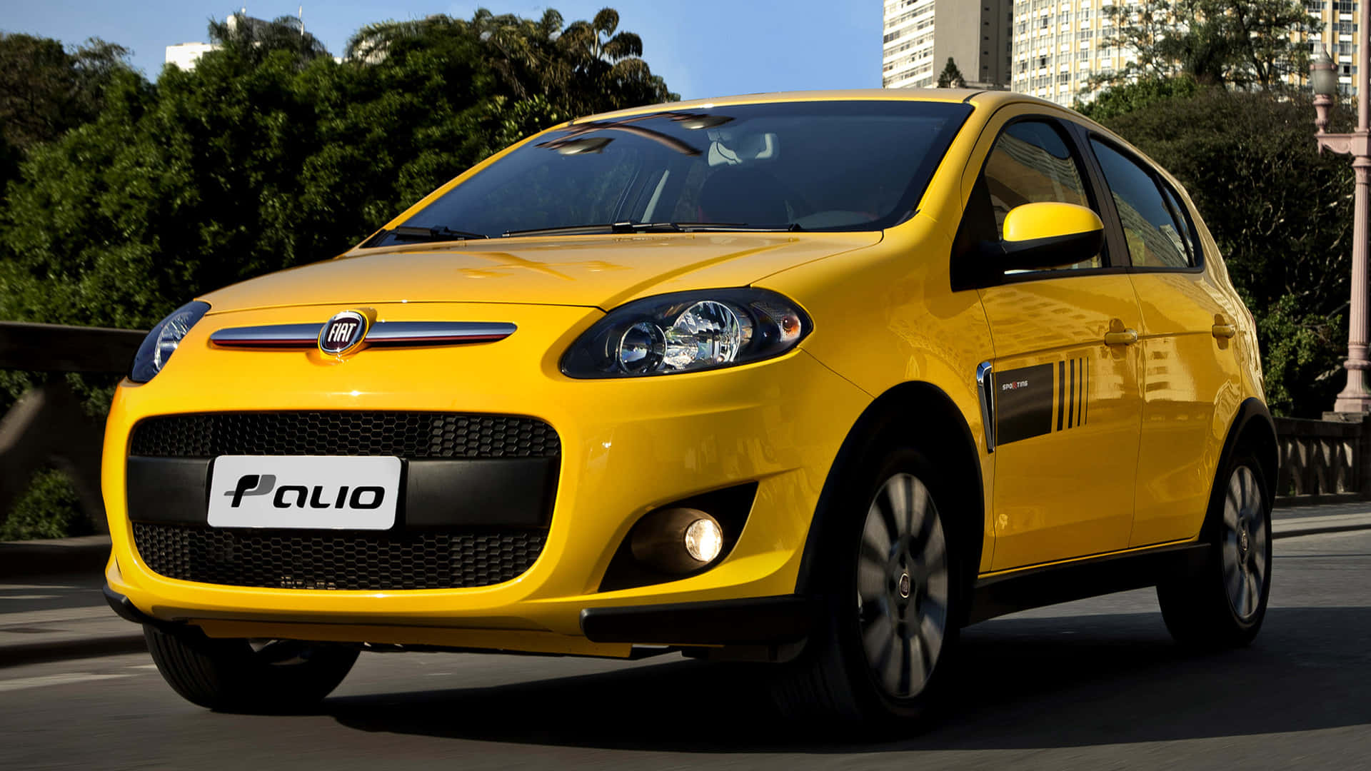 Fiat Palio 桌布 壁紙 — 一輛亮黃色的菲亞特派力奧掀背車停泊在城市的街道上，背景有綠樹和建築物，天空清澈湛藍。