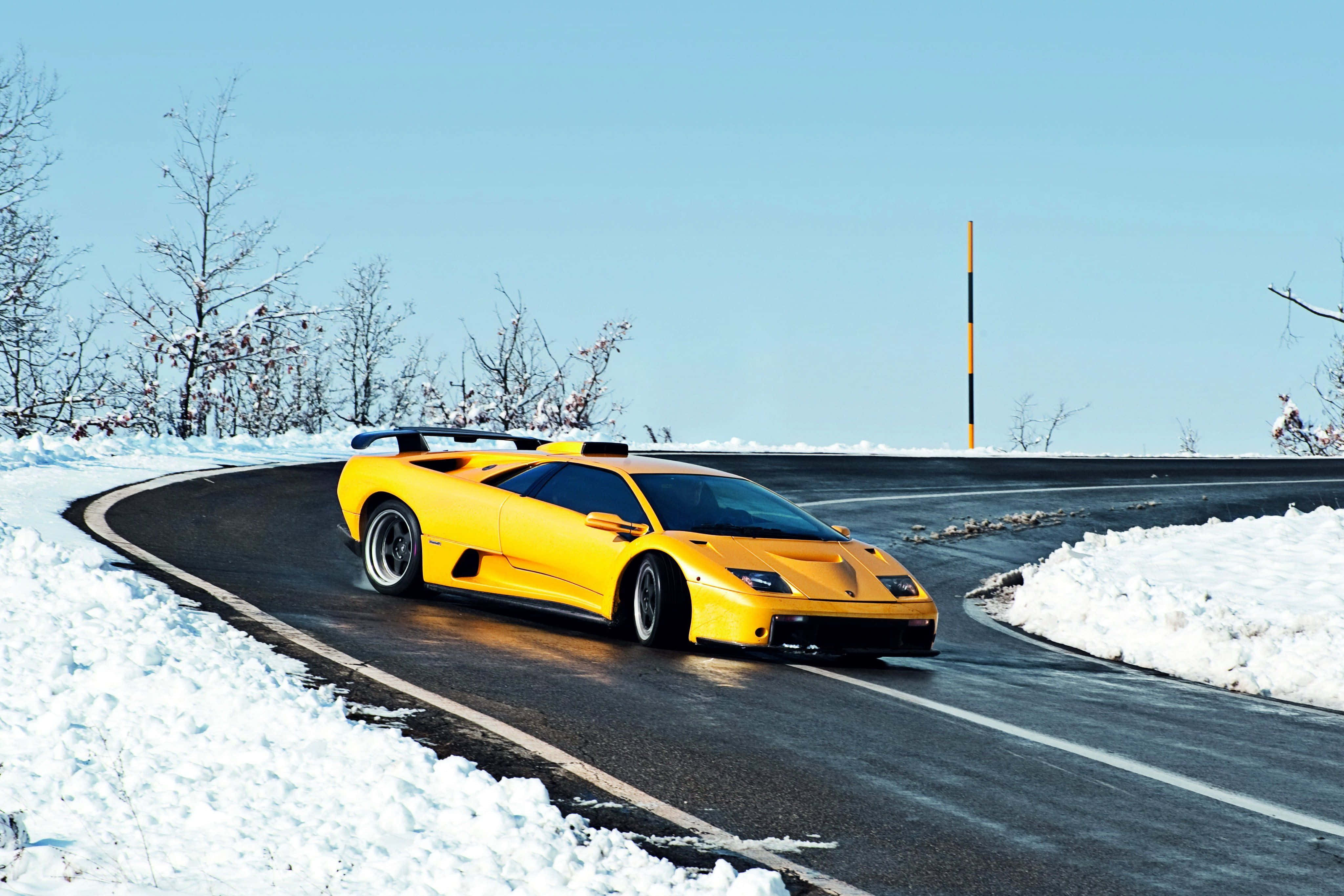 Lamborghini Diablo 桌布 壁紙 — 一輛亮黃色的林寶堅尼 Murciélago 在晴朗的藍天下，沿著積雪的山路奔馳。