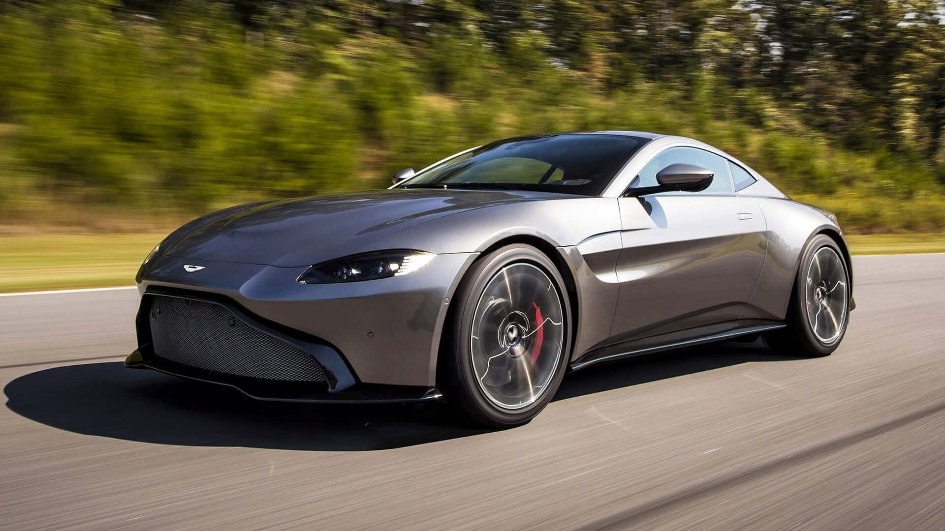 Aston Martin Vantage 桌布 壁紙 — 一輛流線型的銀色Aston Martin Vantage在陽光明媚的道路上疾馳，背景是模糊的綠色樹木。