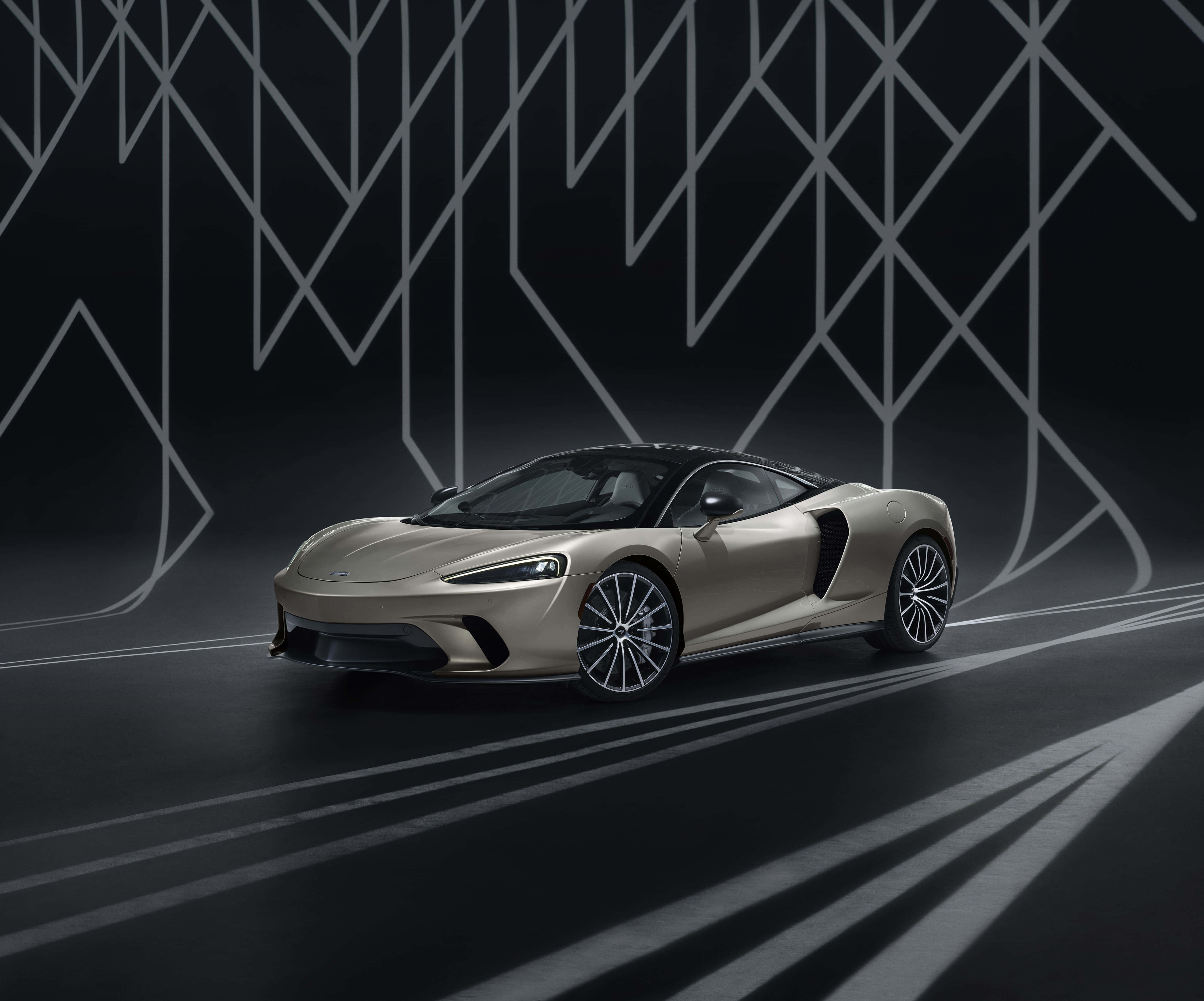 Mclaren Gt 桌布 壁紙 — 一輛流線型金色跑車在帶有幾何白色線條和棱角牆壁圖案的深色工作室地板上滑行。
