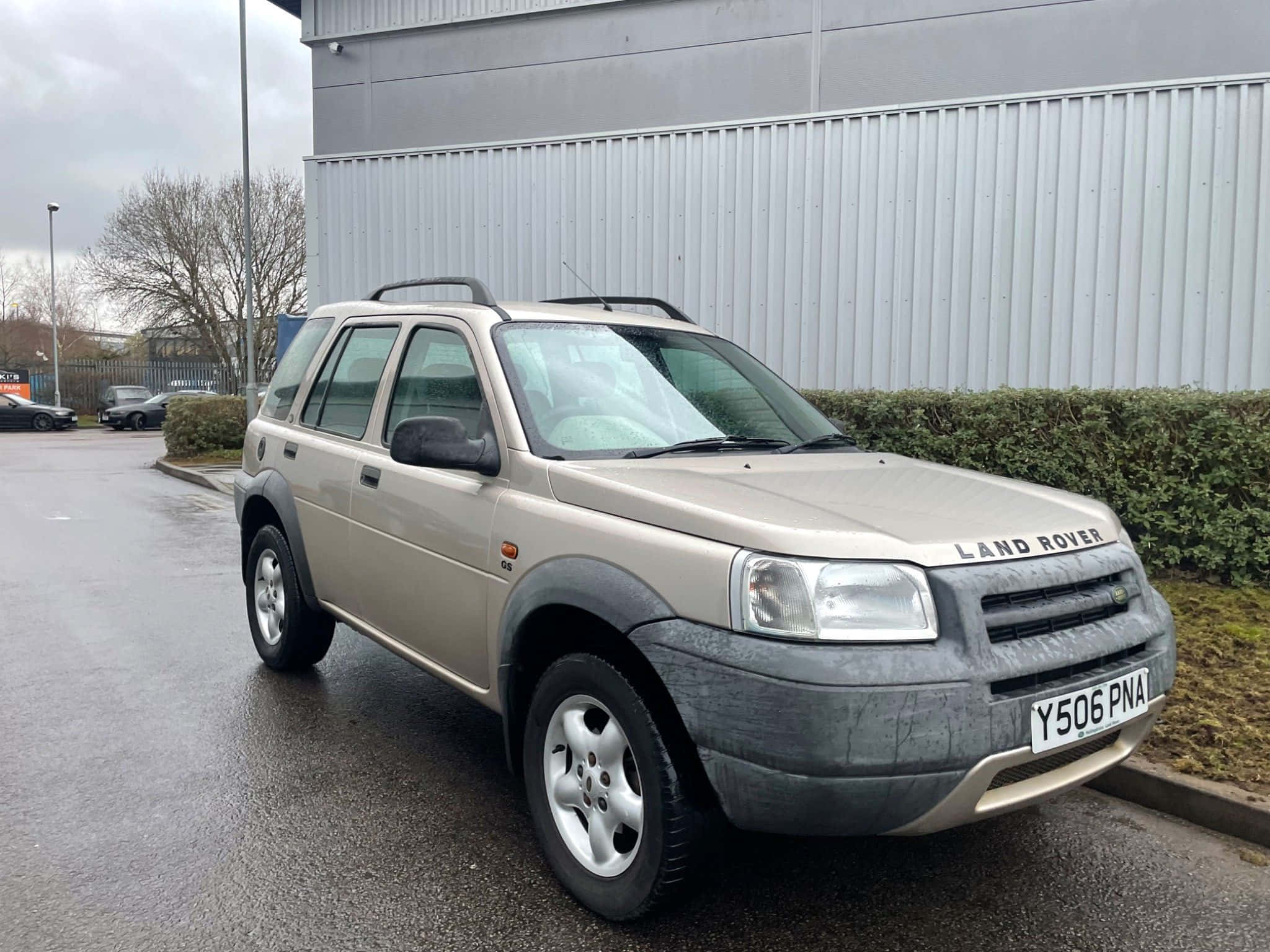 Land Rover Freelander 桌布 壁紙 — 一輛米色Land Rover Freelander停泊在潮濕的瀝青停車場，旁邊有樹籬和工業建築，天空多雲。